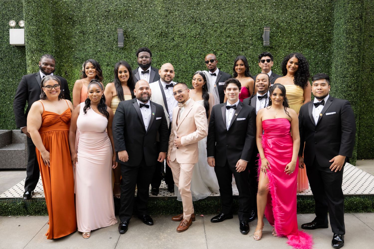 Wedding party poses outdoors against a green hedge wall wearing formal attire in black and bright colors.
