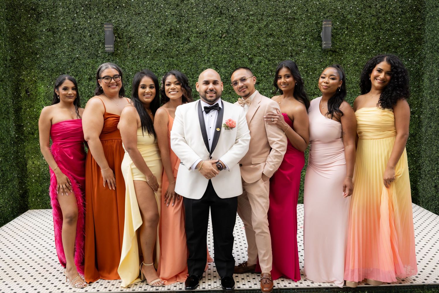 Wedding party stands in a line wearing dresses in shades of rust, pink, and yellow against greenery backdrop.