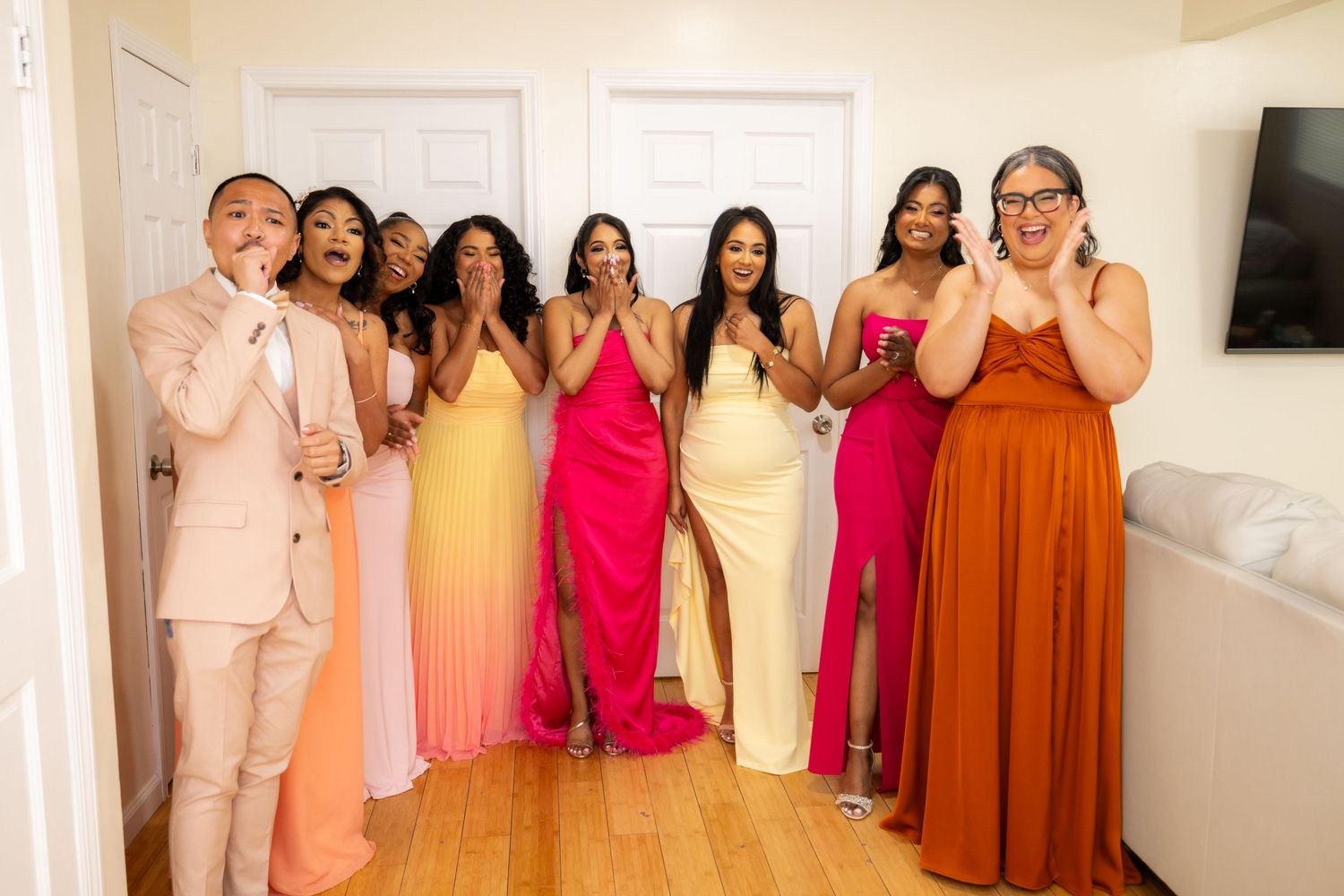 A group of people pose together in a hallway wearing coordinated formal attire in shades of pink, red and orange.