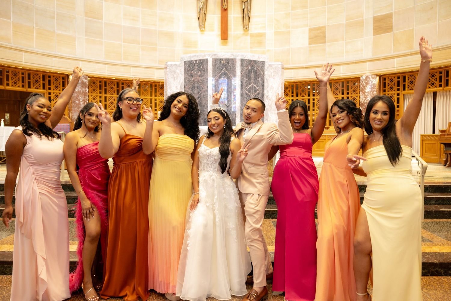 Wedding party in colorful dresses standing together in elegant indoor venue with ornate architecture.
