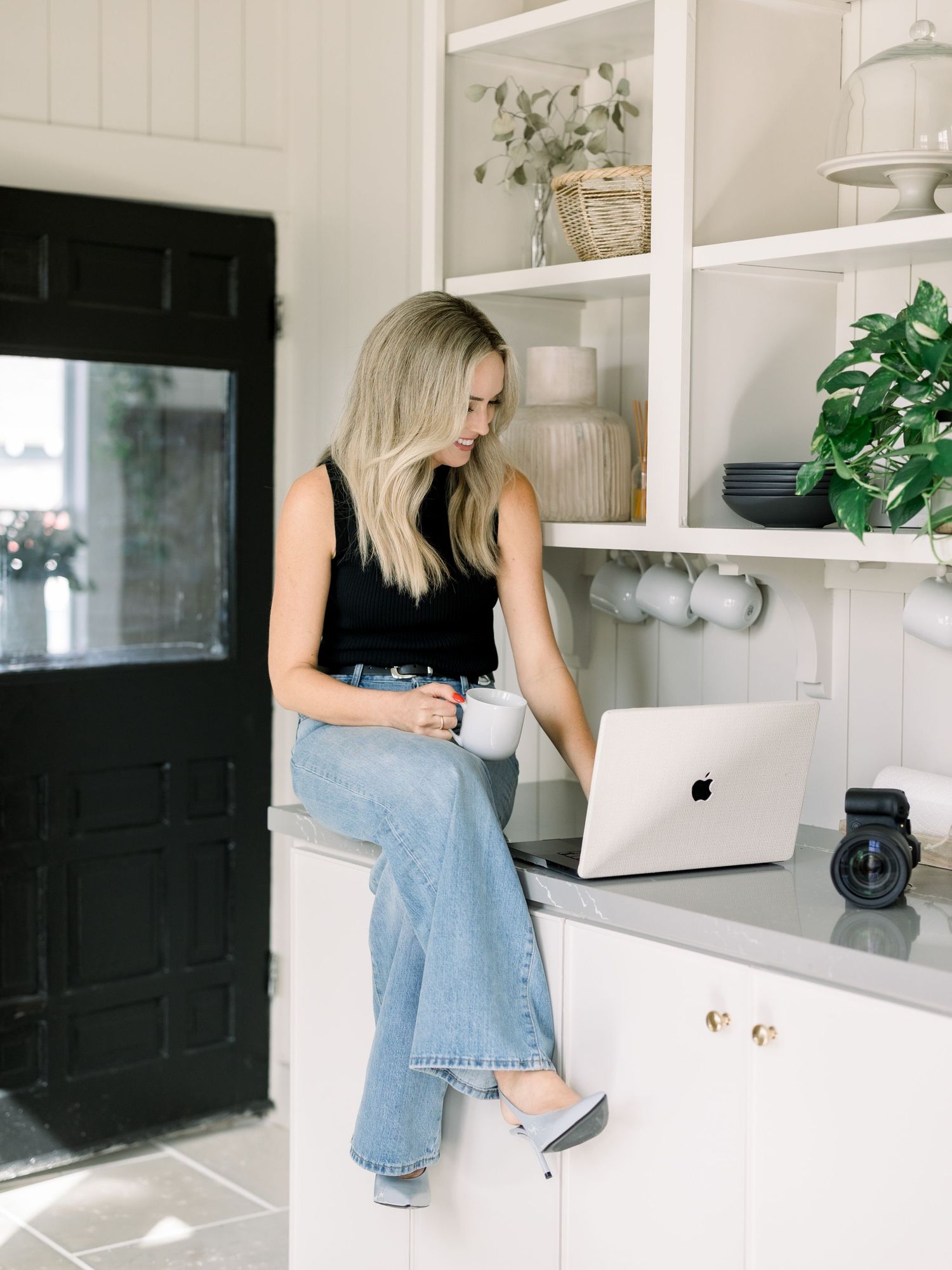 Blonde woman sitting on kitchen counter with MacBook and coffee mug in bright white modern kitchen.