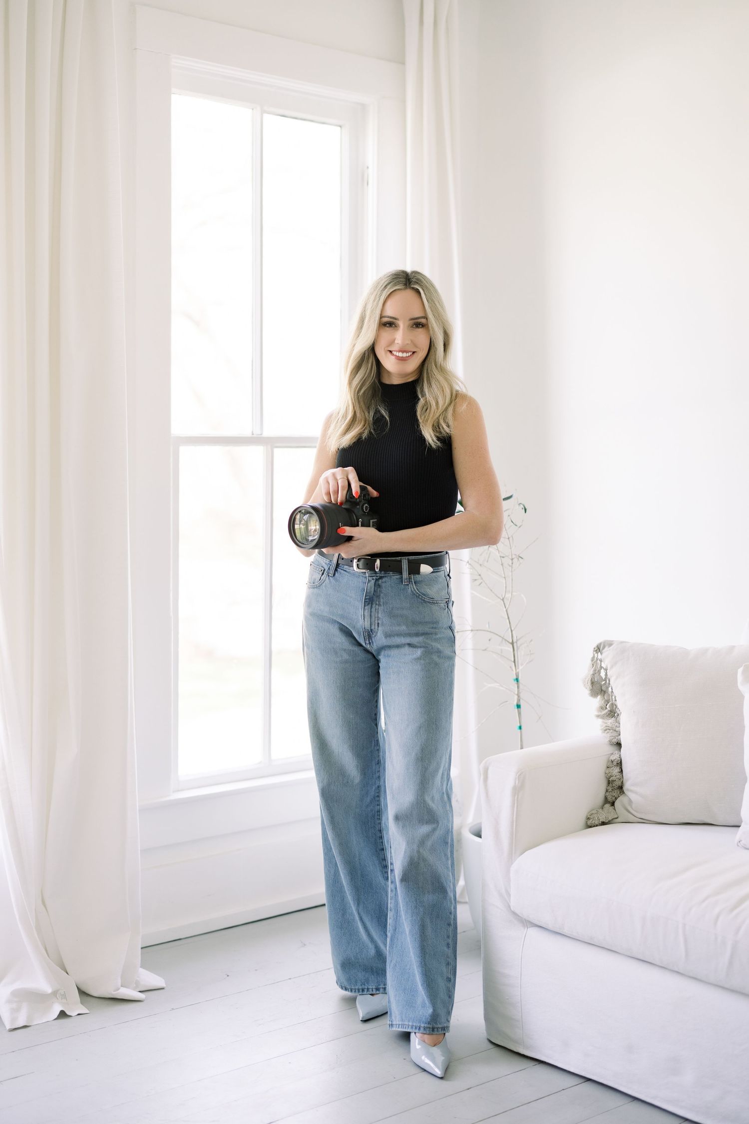 Blonde woman in black sleeveless top and wide-leg jeans holding a camera, standing in a bright white room.
