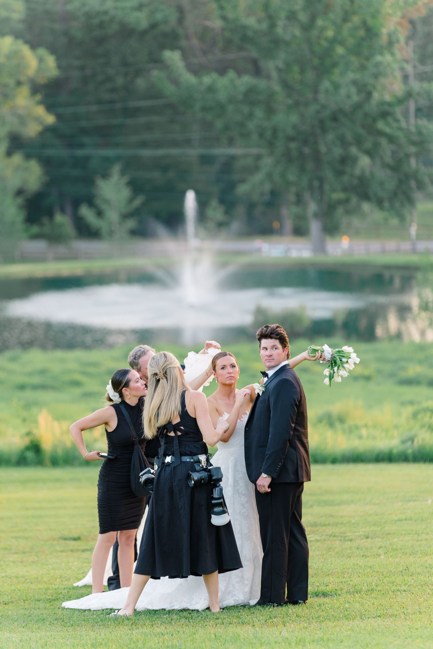 Wedding party posing outdoors near a fountain, with bridesmaids in black dresses and bride in white gown on green lawn.