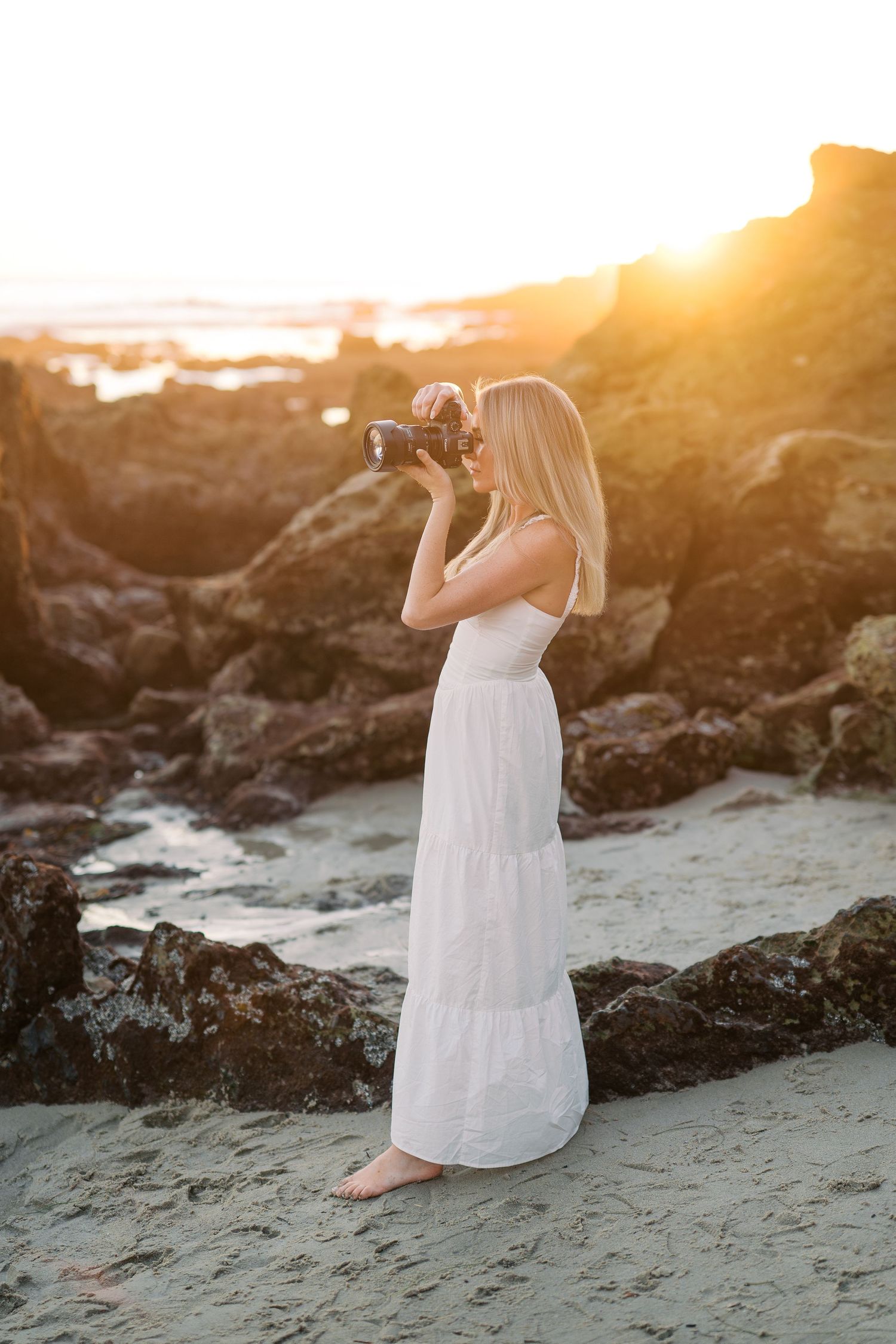 Blonde woman in white maxi dress photographing rocky beach at golden sunset, standing barefoot on sand.