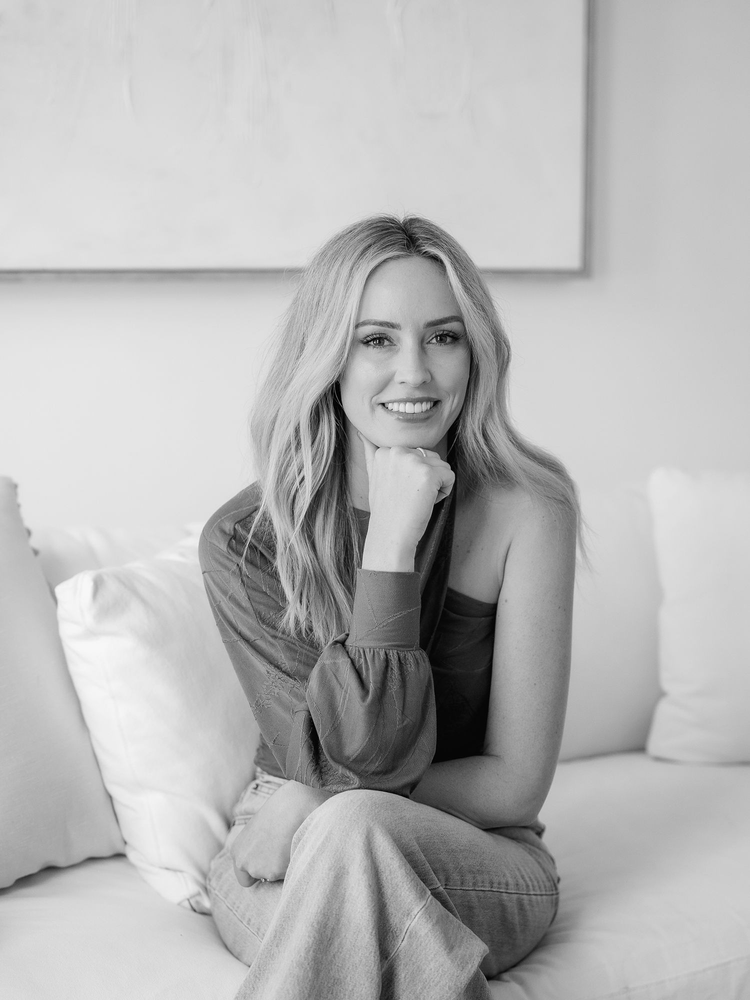Black and white portrait of a smiling blonde woman sitting on a white sofa in a bright modern interior.