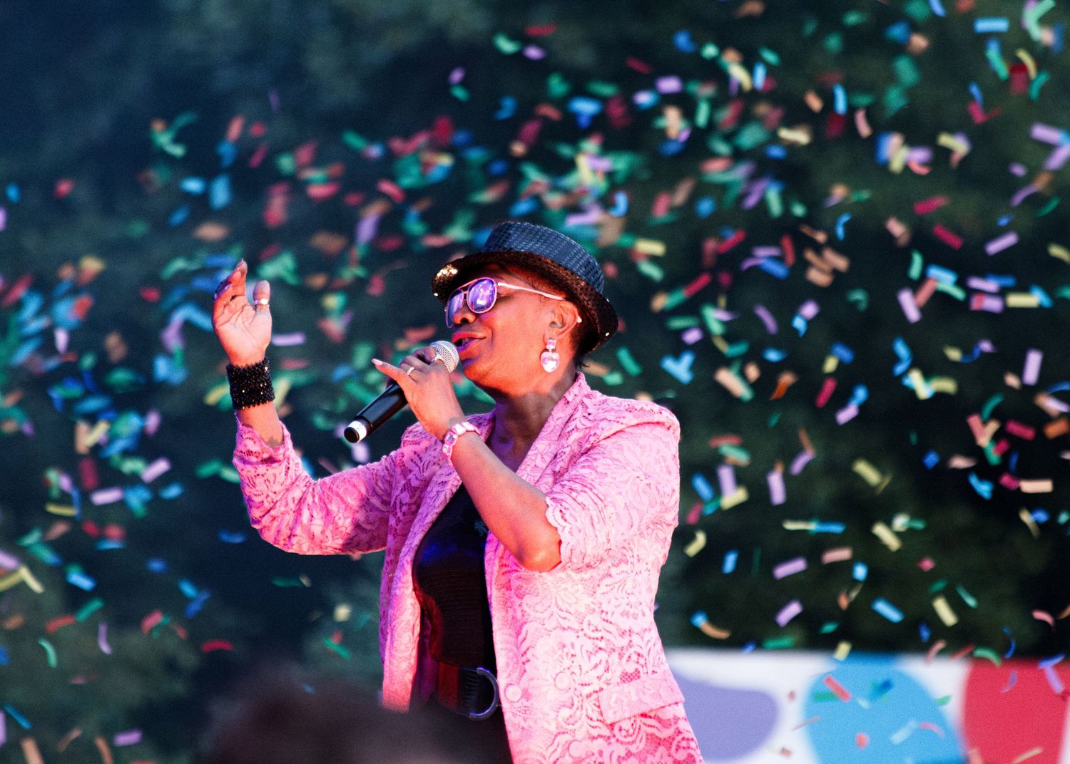 A performer in a pink sequined jacket and black hat sings on stage while confetti falls in vibrant colors around them.