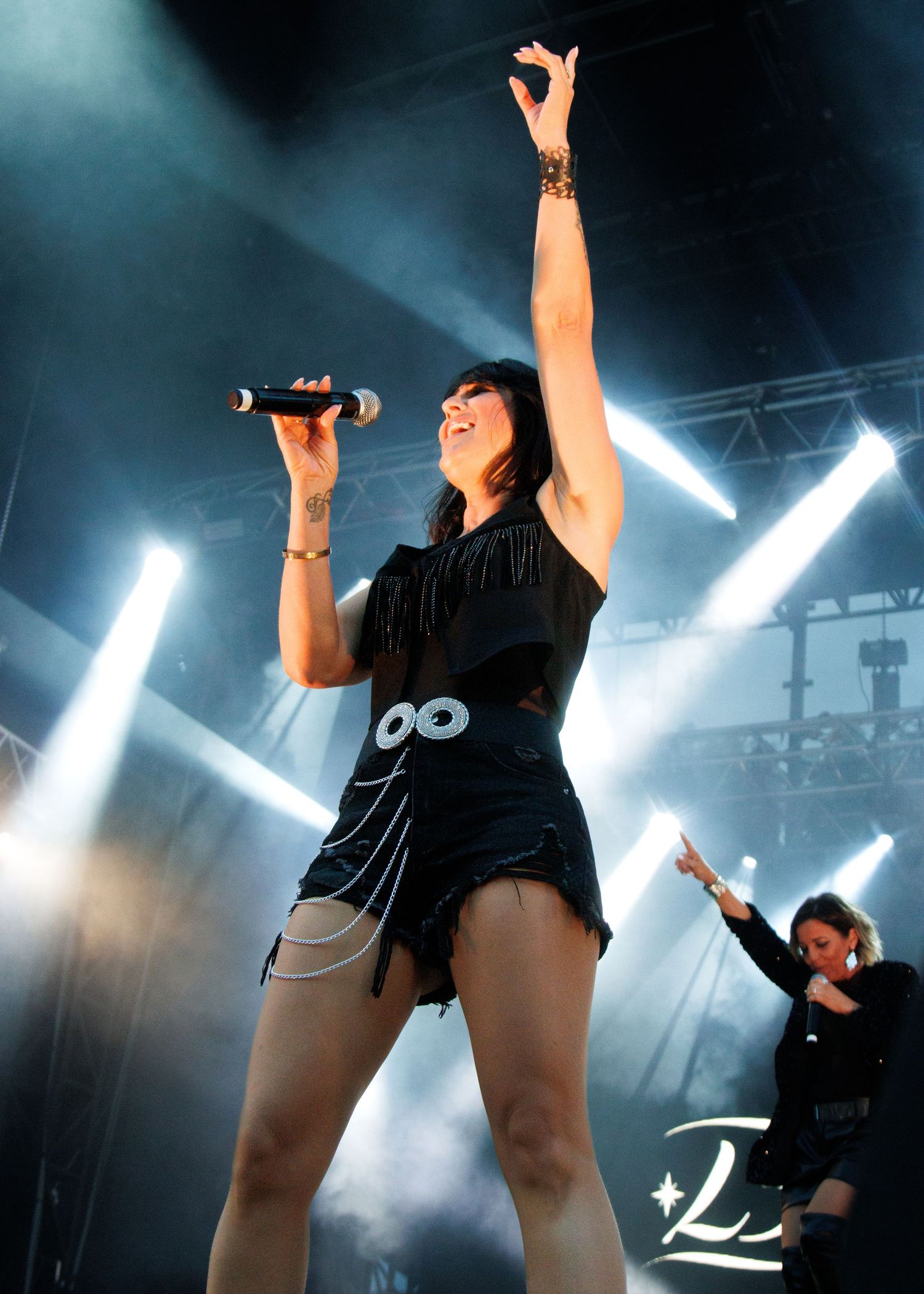 A performer in a black outfit sings on stage with dramatic blue lighting and raised arm during an energetic concert performance.