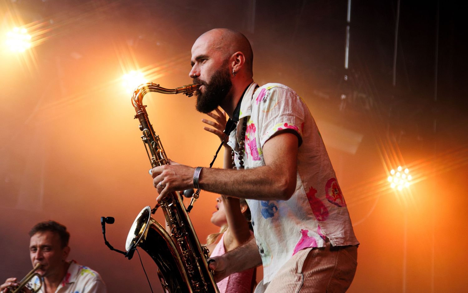 Saxophone player performs energetically on stage with warm orange lighting during a live concert performance.