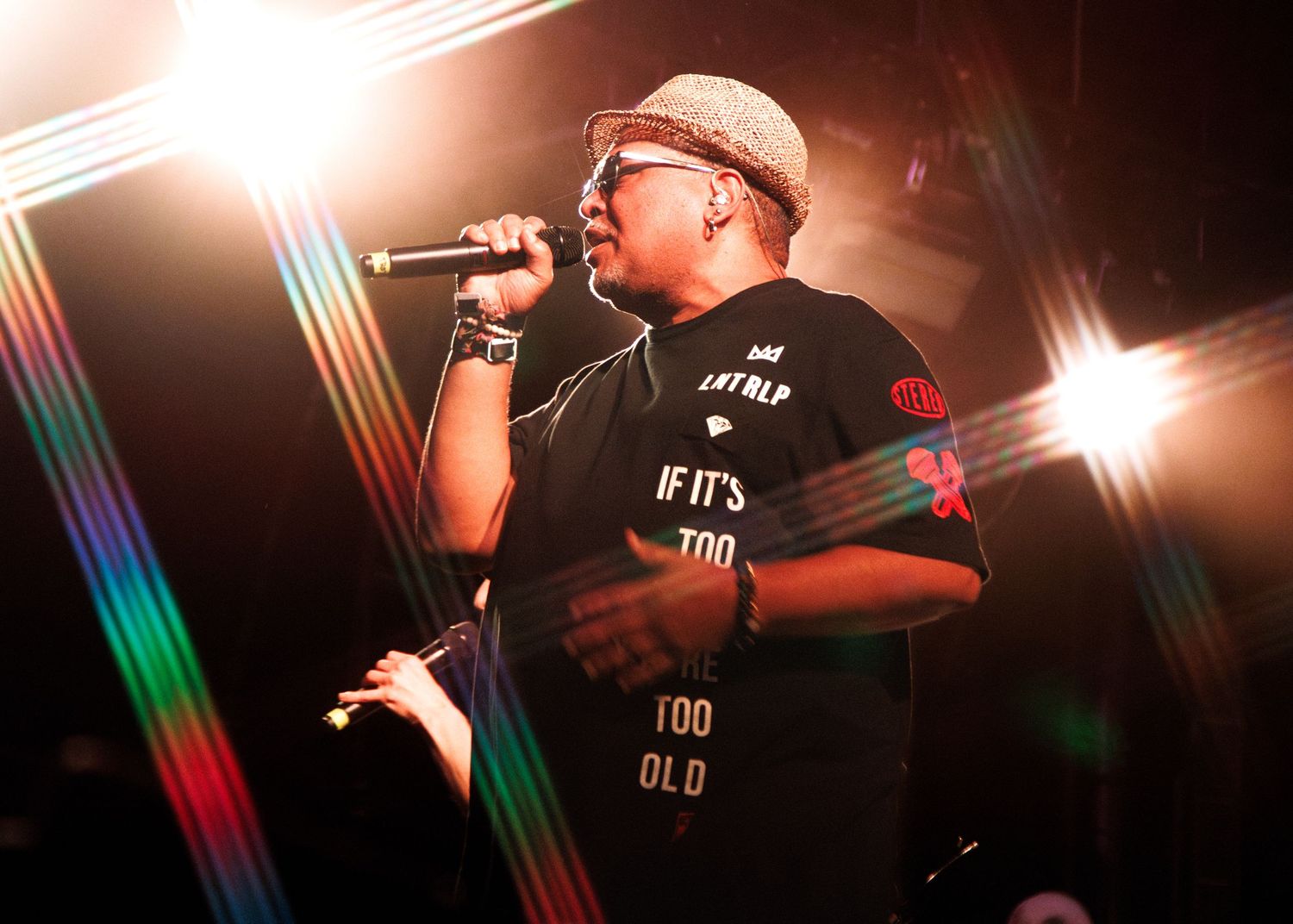 Hip hop artist performs on stage with dramatic lighting and colorful beams in the background during live concert.