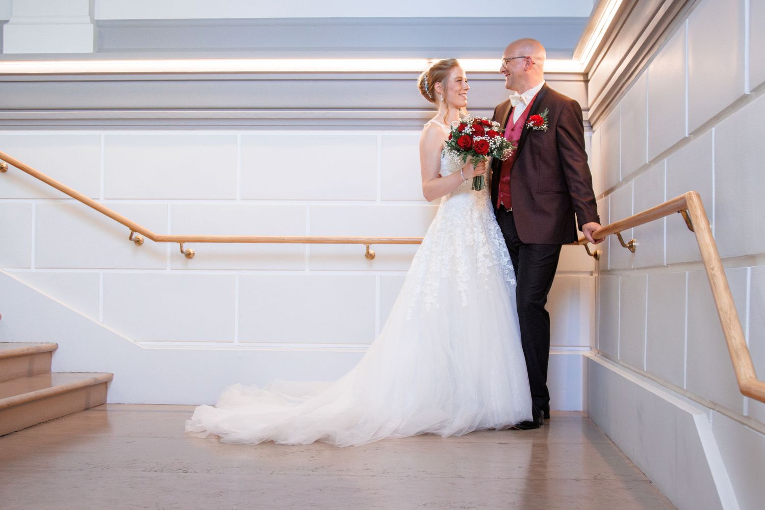 A bride in a white gown and groom in burgundy suit pose on a staircase with red roses.