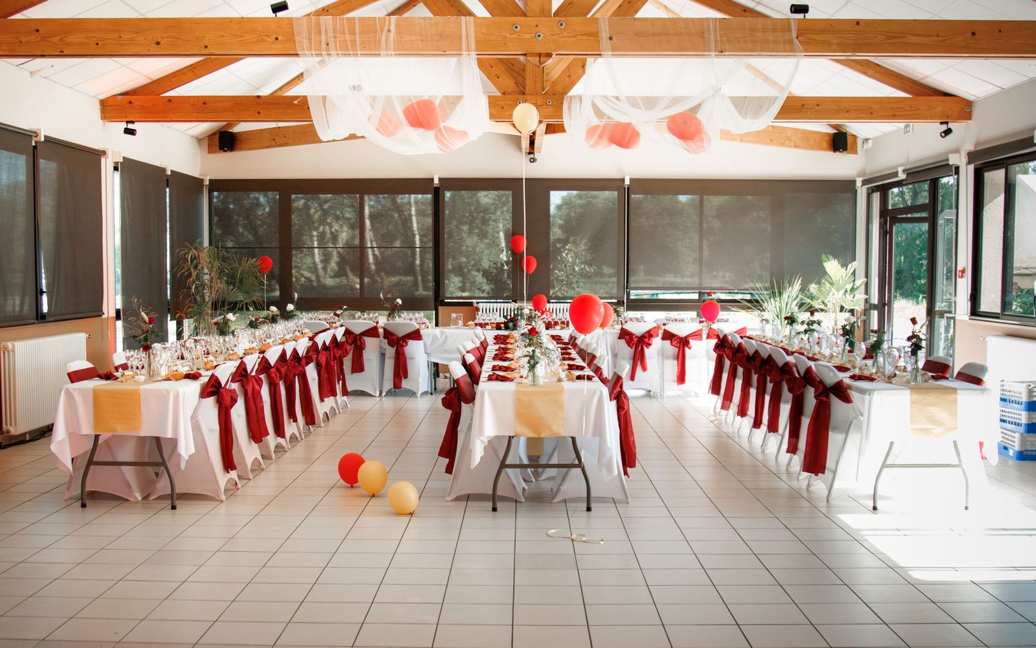 Decorated wedding reception hall with red and white balloons, long tables, and red chair sashes.