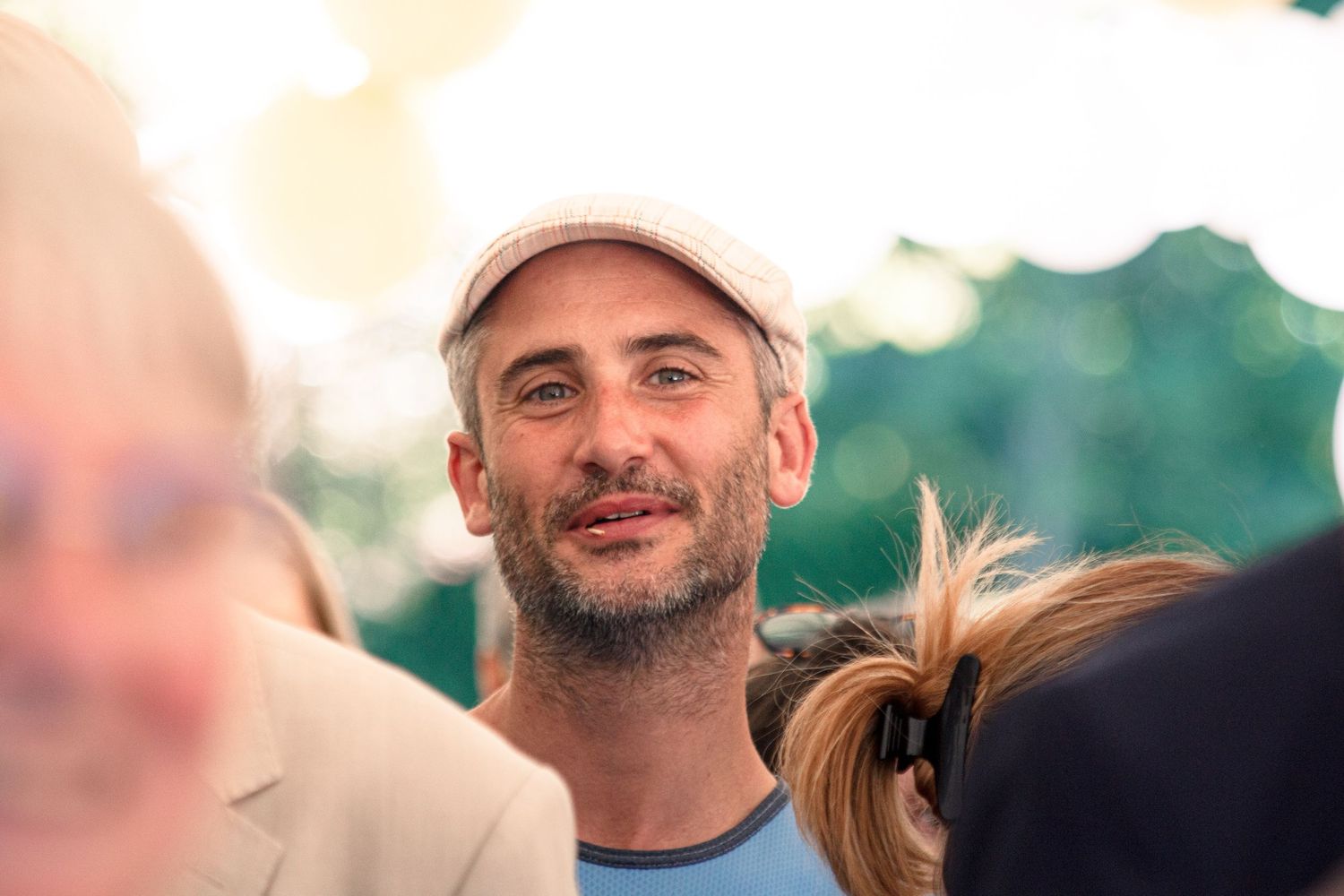 Smiling man wearing a flat cap and blue shirt enjoying an outdoor wedding reception in warm sunlight.