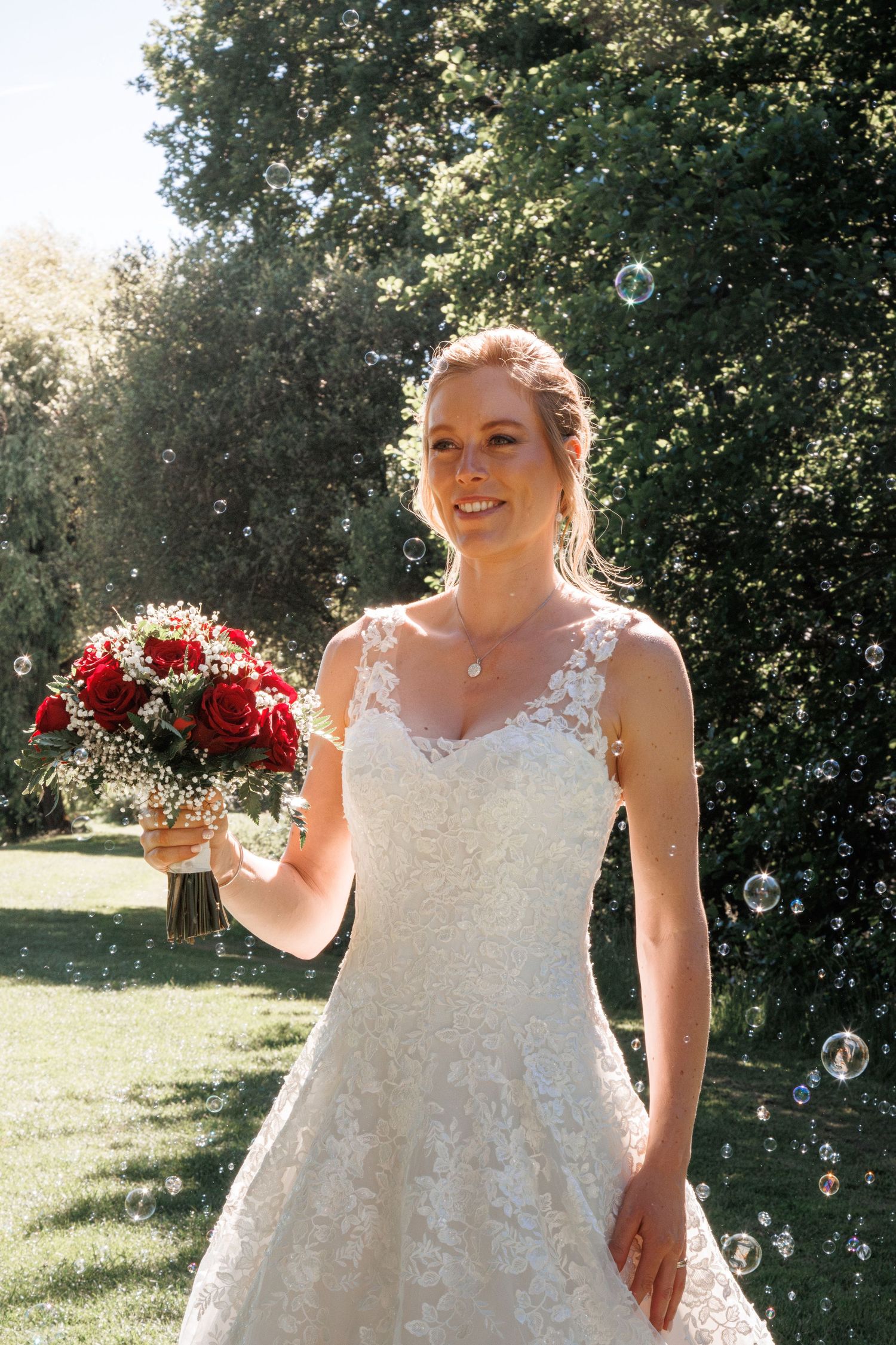 Radiant bride in lace A-line wedding gown holds red roses bouquet surrounded by floating bubbles outdoors.