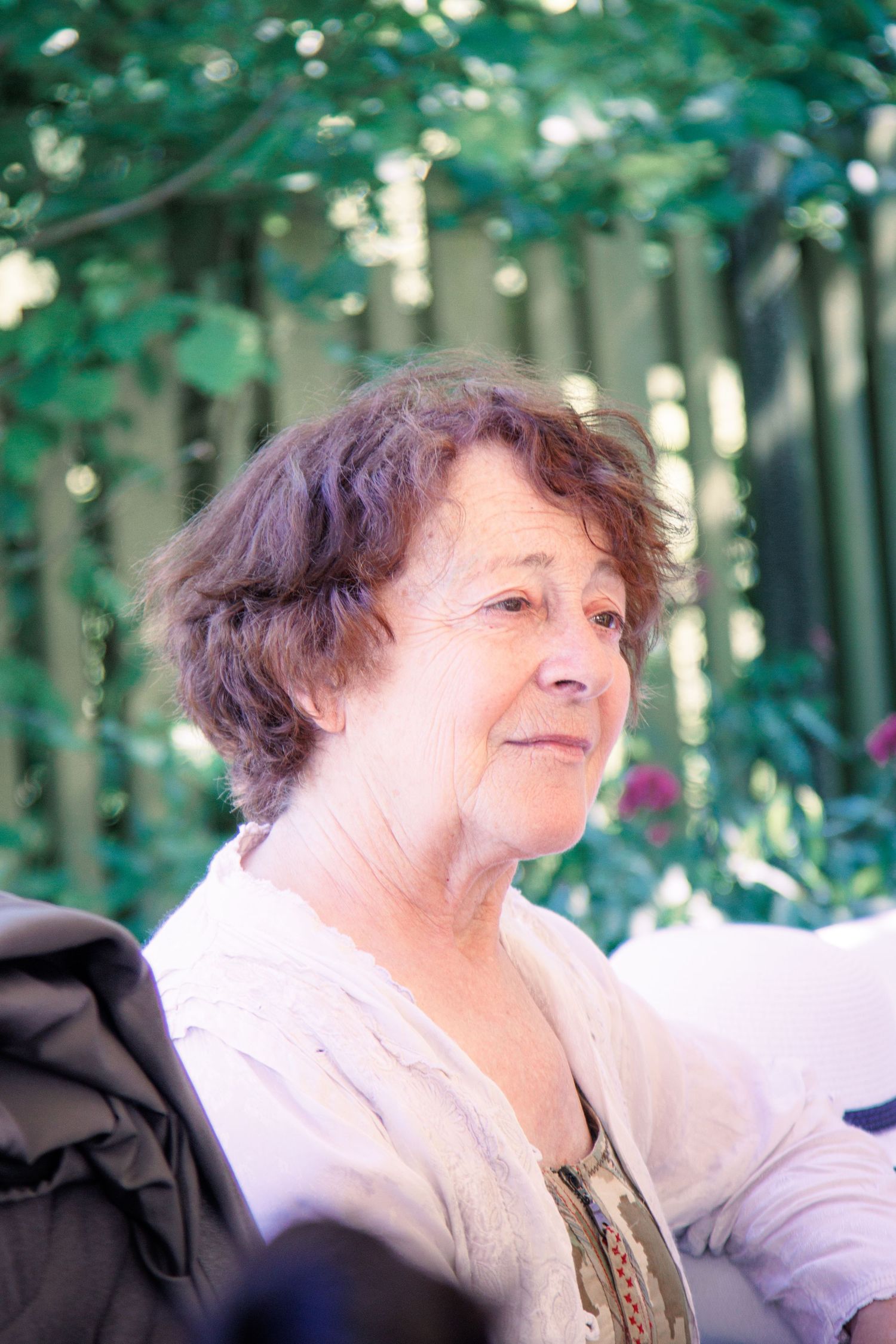Elegant older woman with short brown hair seated outdoors at a wedding, surrounded by lush green foliage.