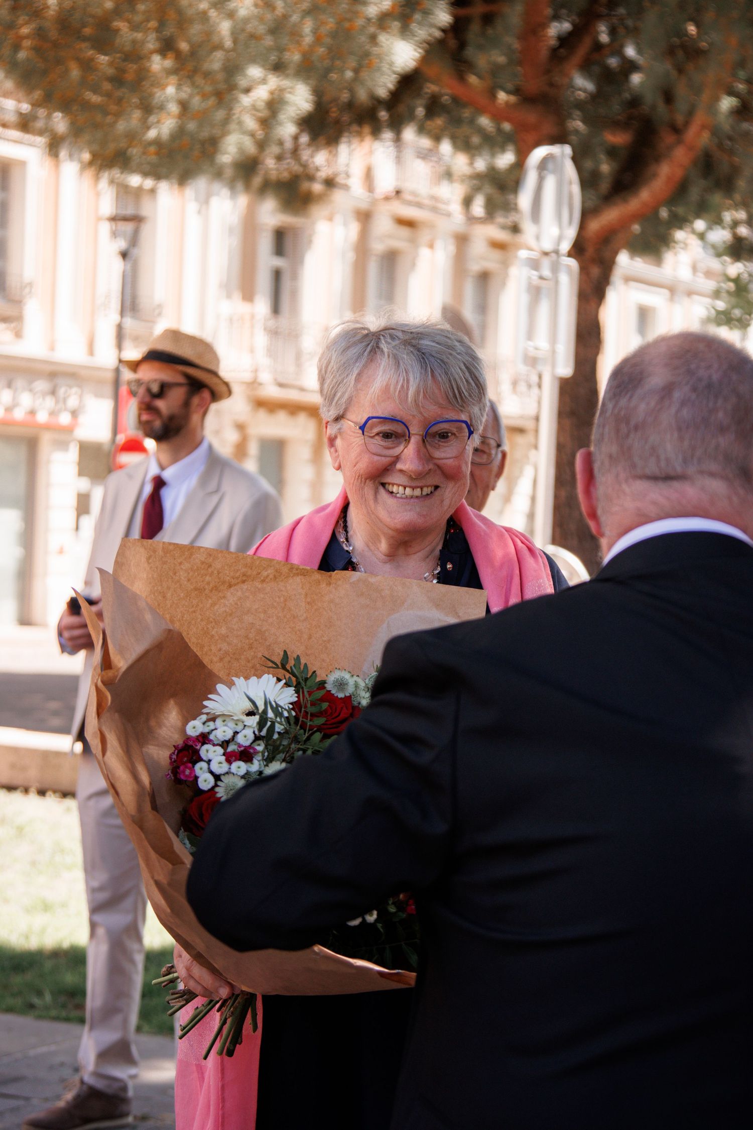 Smiling elderly woman with glasses receiving a bouquet of flowers outdoors at a wedding celebration.