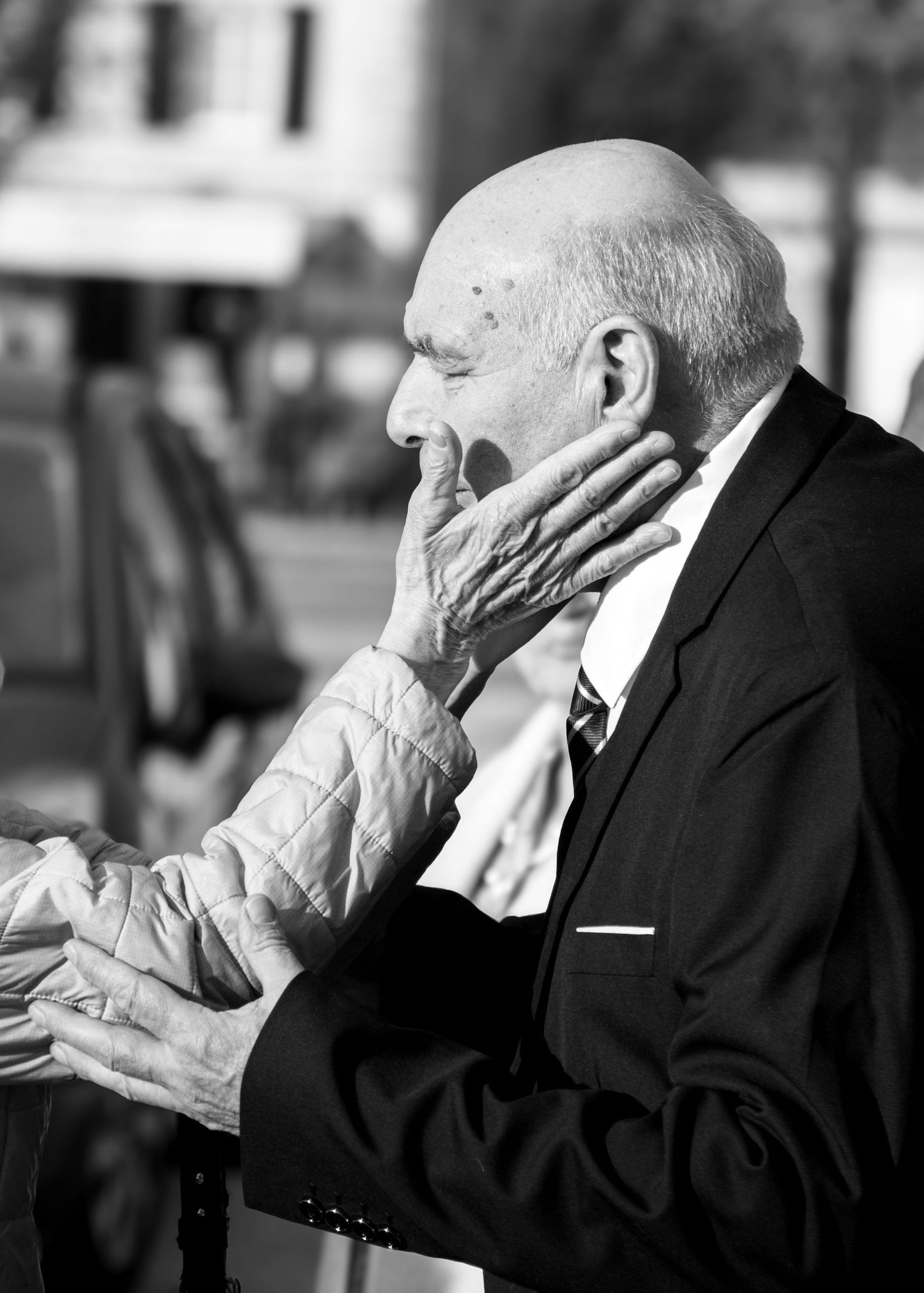 Elderly man in suit tenderly touched on face by wrinkled hands in black and white portrait photo.