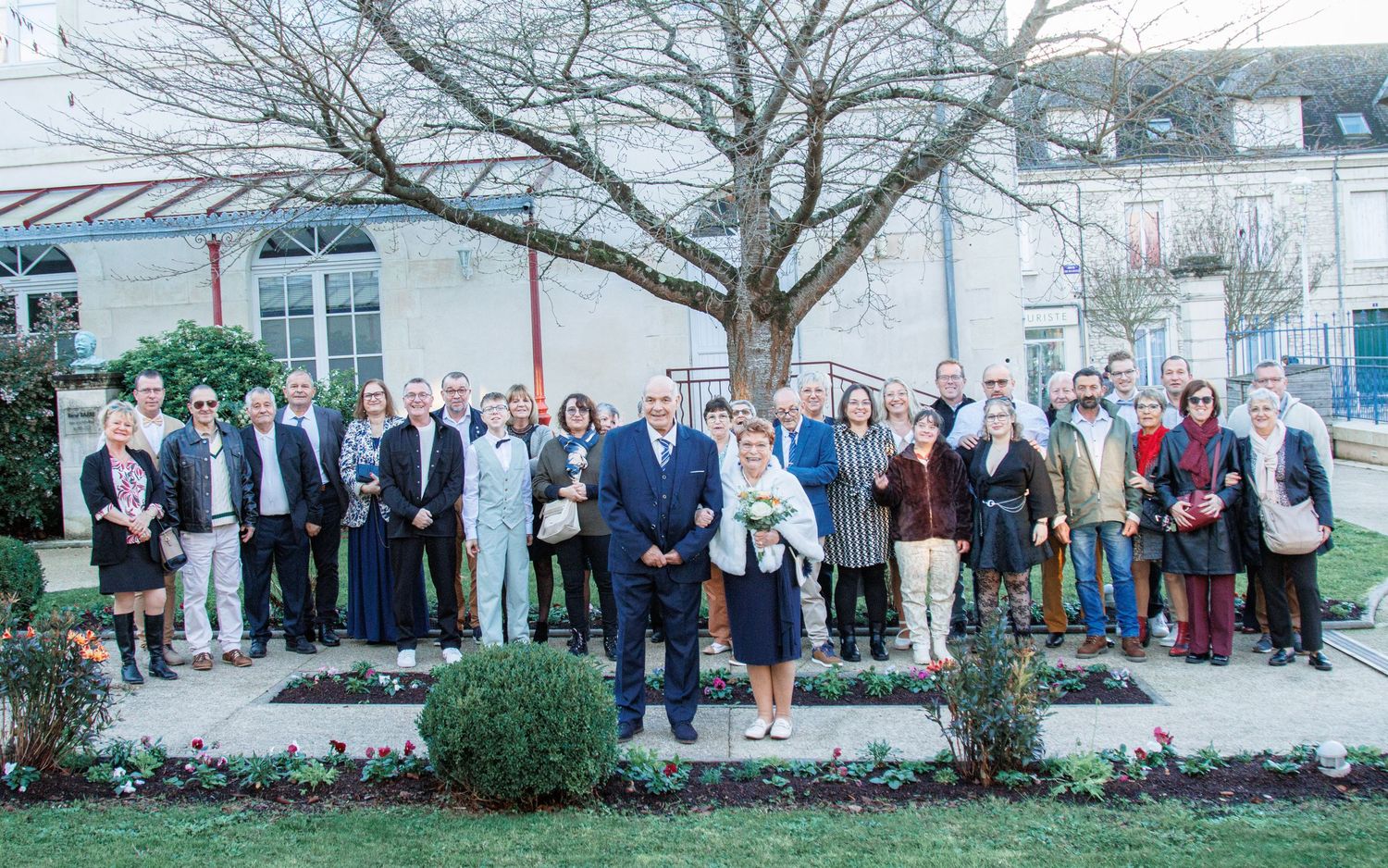 Large group of professionals posing outdoors in a formal garden setting with bare trees and building in background.
