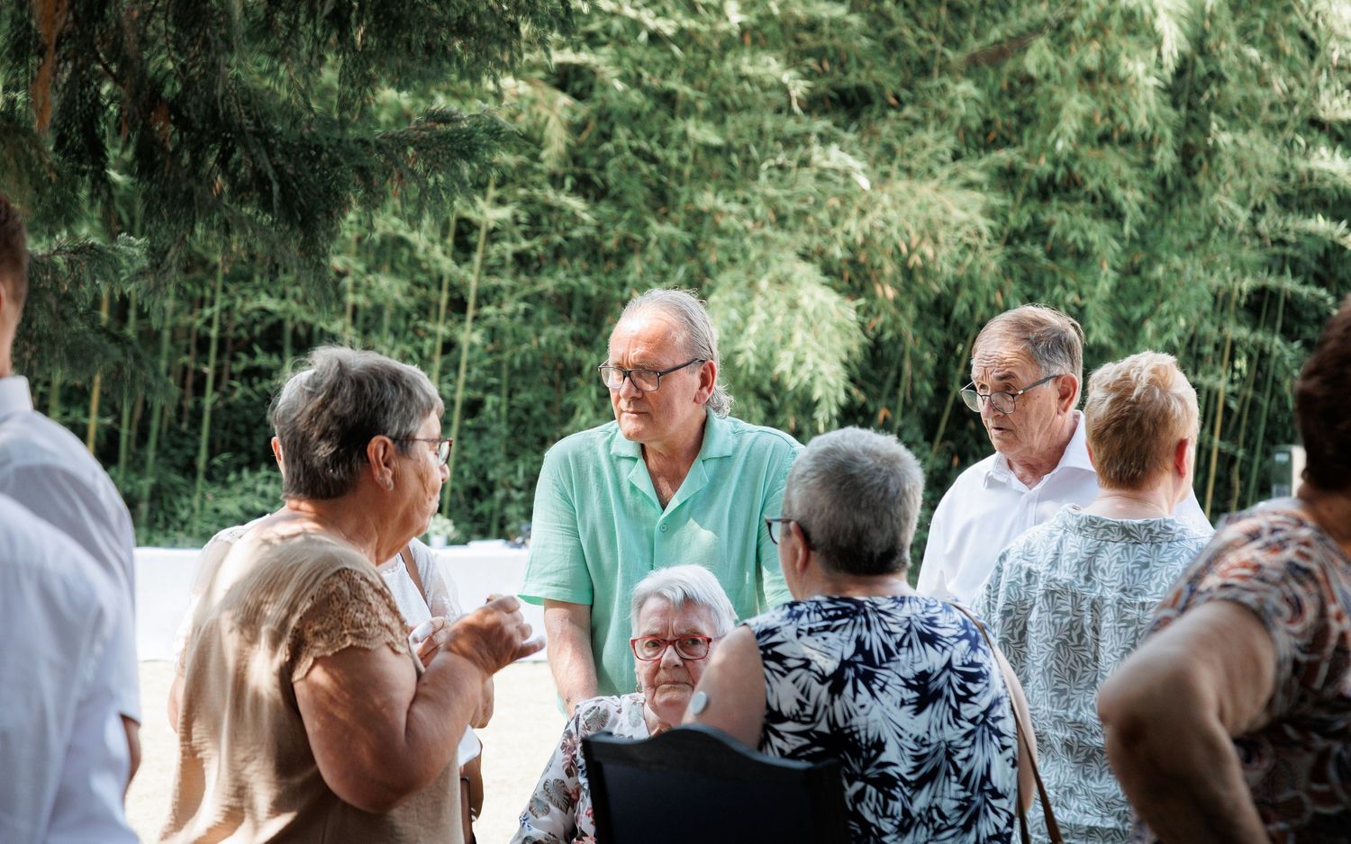Elderly wedding guests gather and converse outdoors surrounded by lush green bamboo trees.