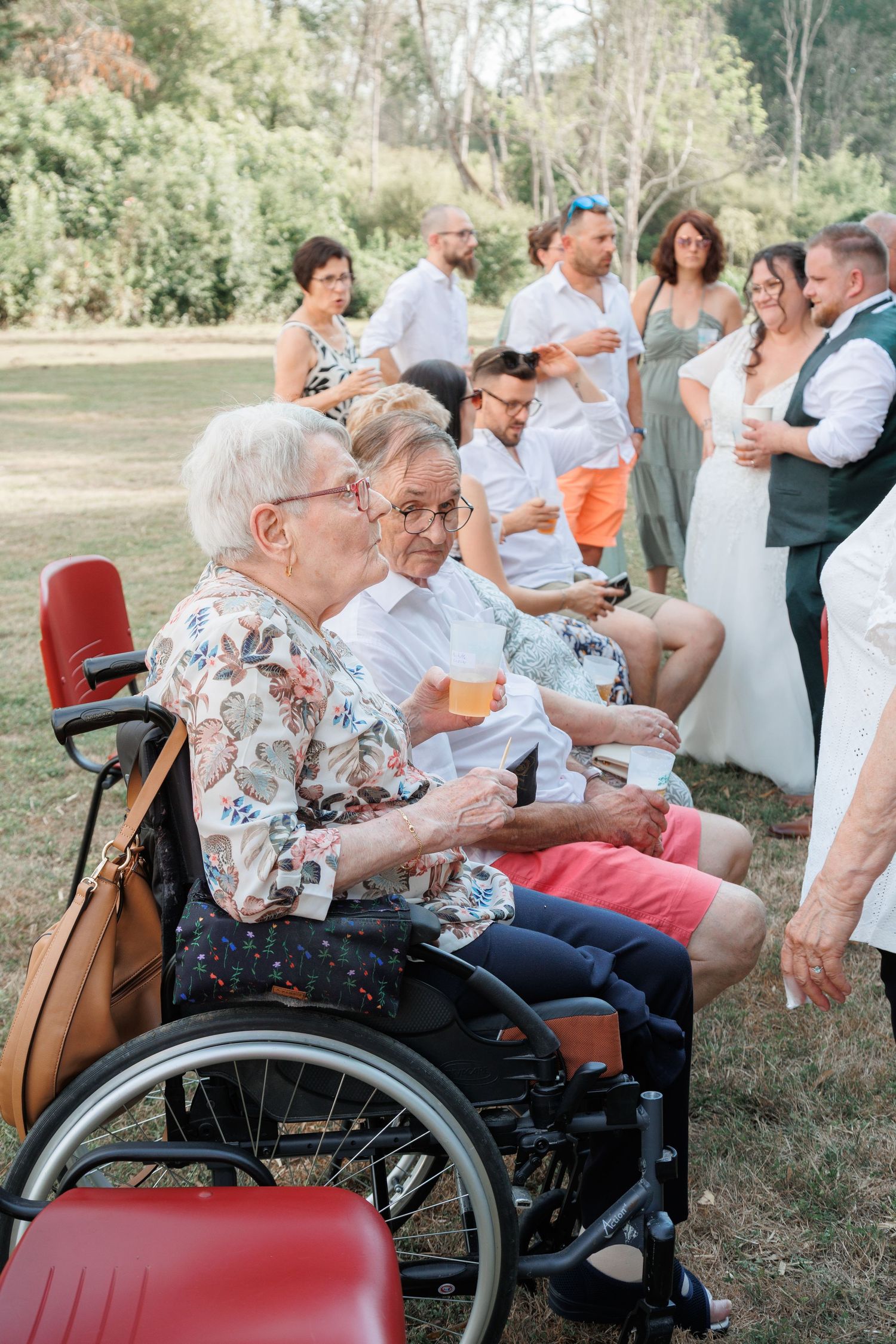 Elderly couple in wheelchairs enjoying outdoor wedding reception with guests mingling in background.