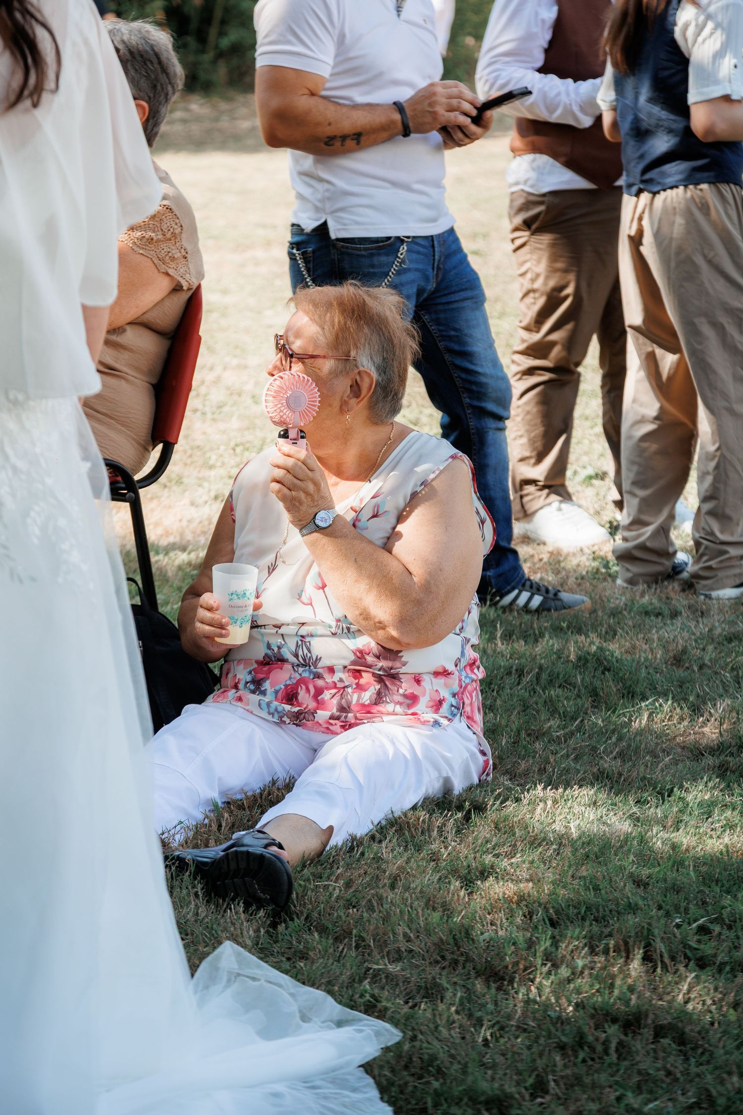 Elderly woman sitting on grass holding pink lollipop at outdoor wedding.