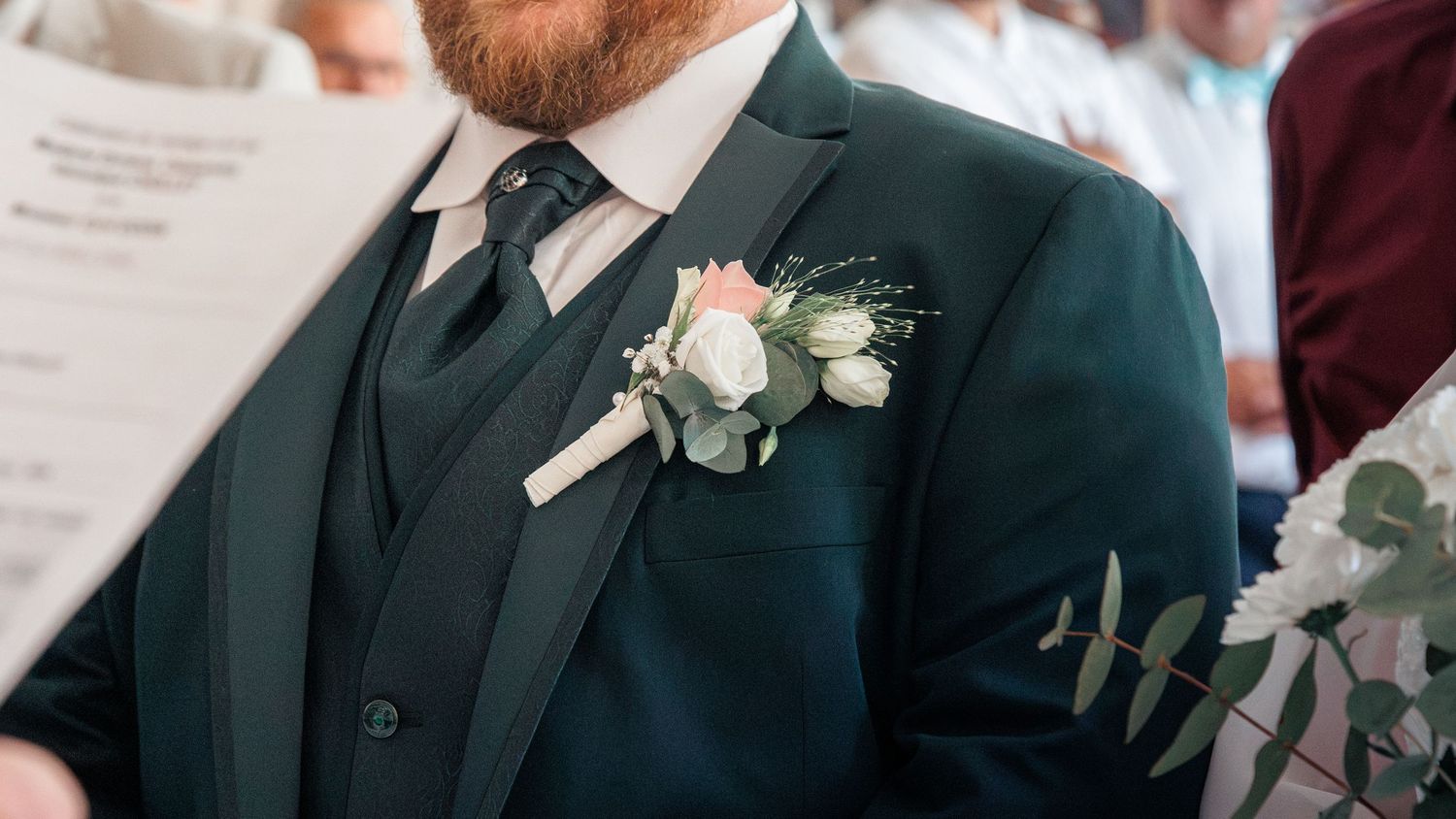 Groom in a dark teal three-piece suit with a white rose boutonniere at a wedding ceremony.