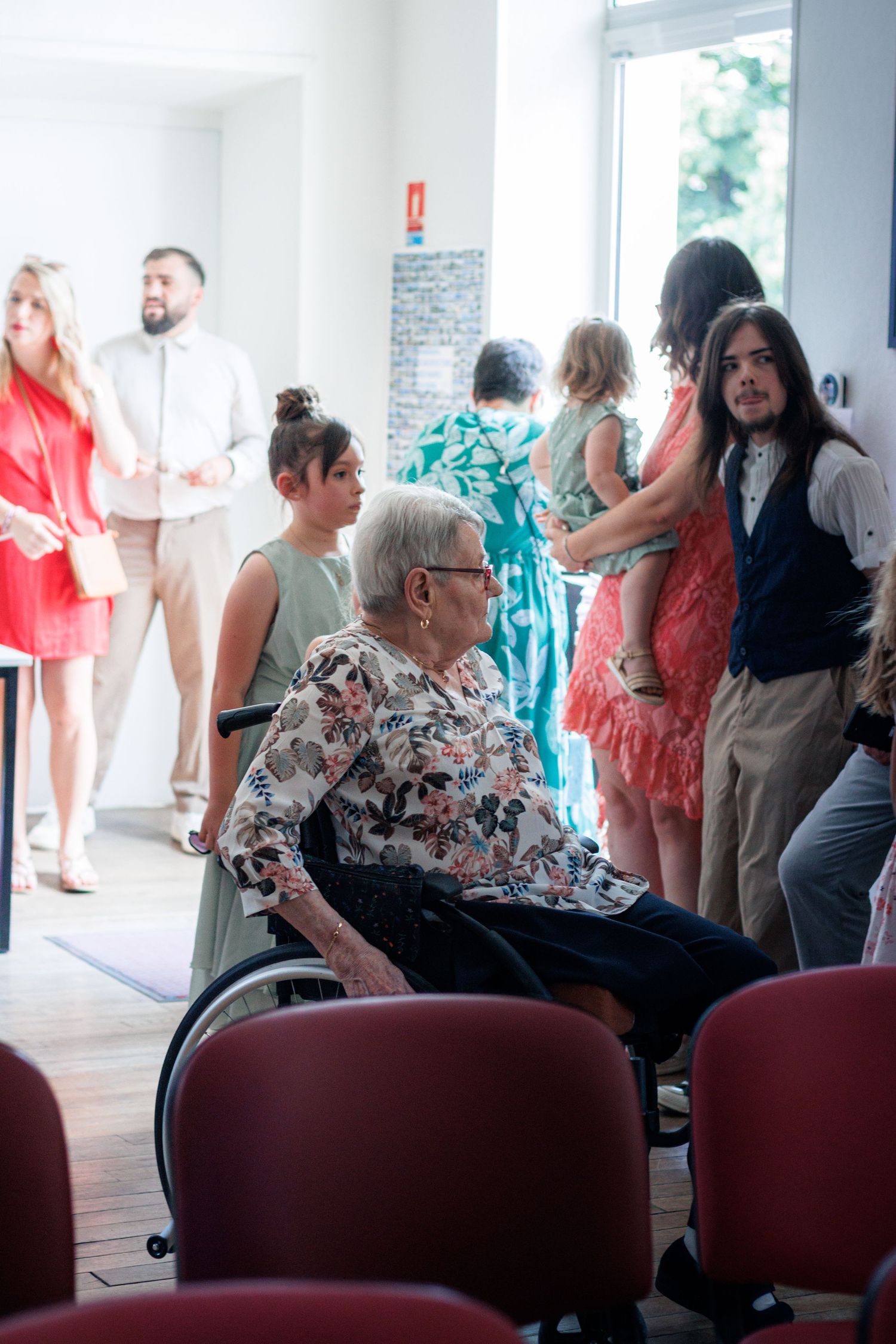 Elderly woman in wheelchair at wedding ceremony surrounded by guests in colorful formal attire.