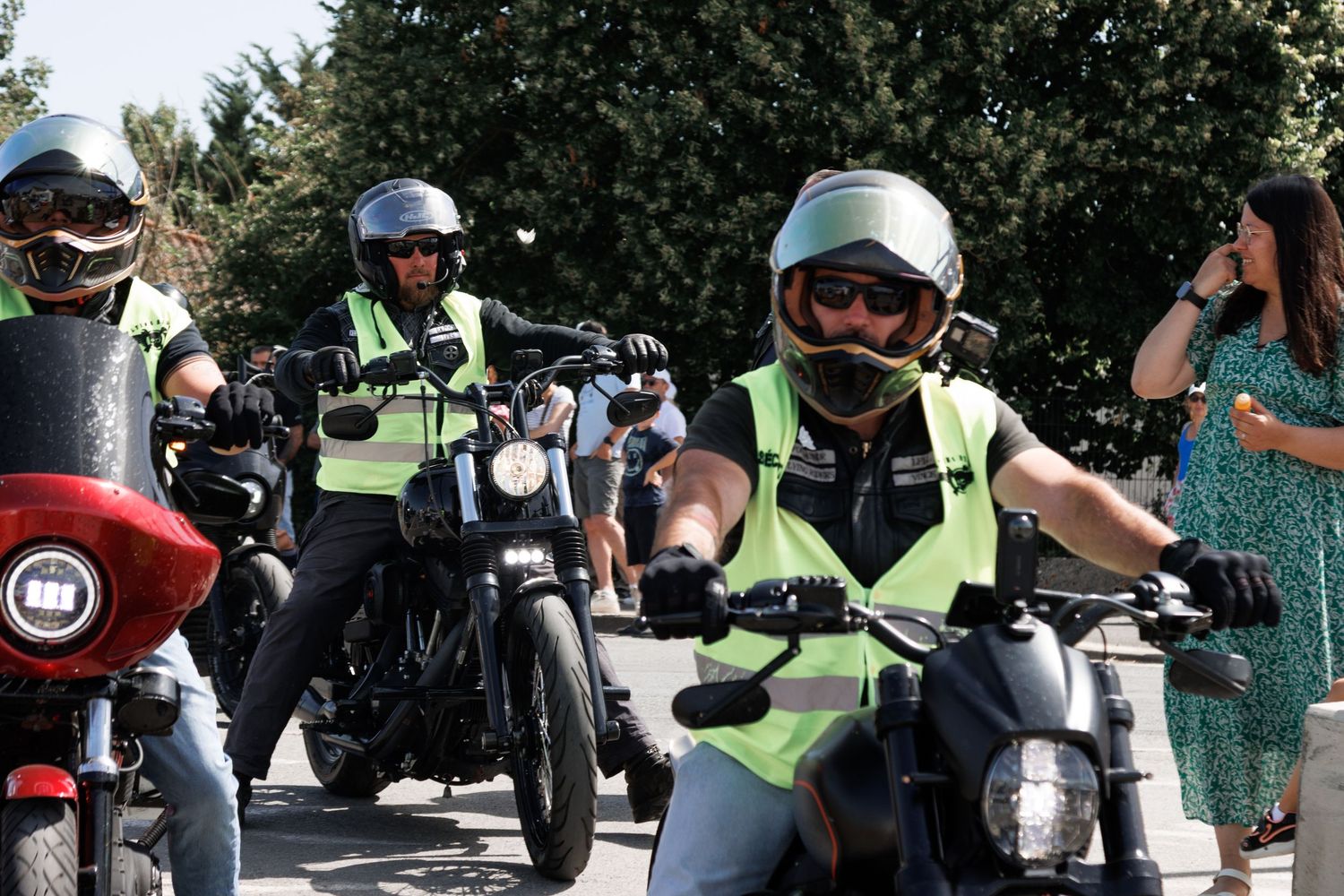 Two motorcycle riders wearing helmets and green safety vests ride motorcycles on a sunny street.