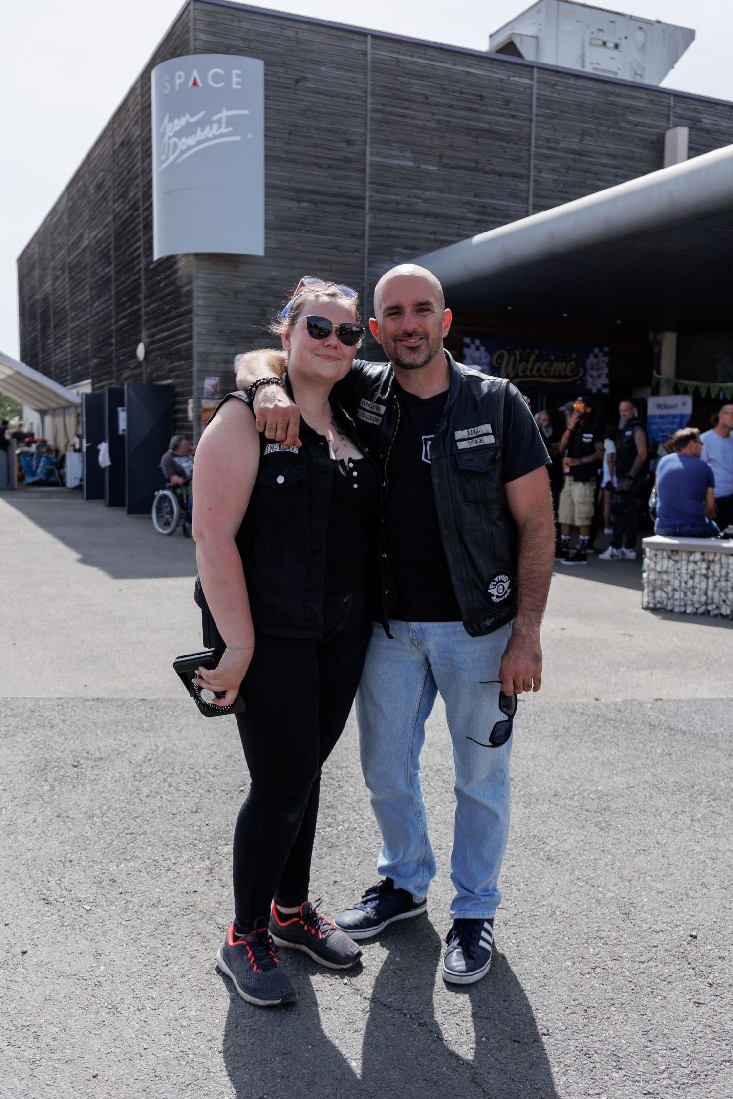 A couple posing together outdoors in front of a modern building, woman in black outfit and man in biker vest.