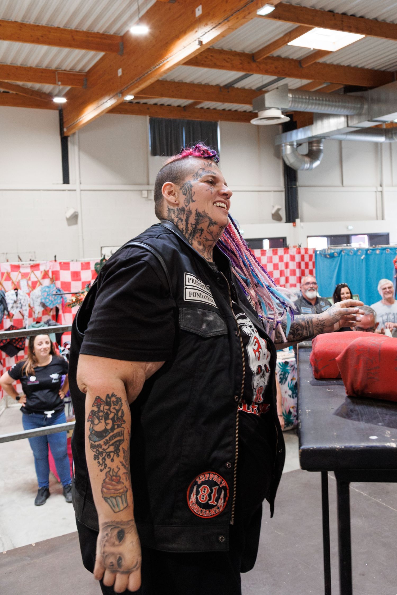 Heavily tattooed person with pink braids wearing a black biker vest at an indoor tattoo convention.