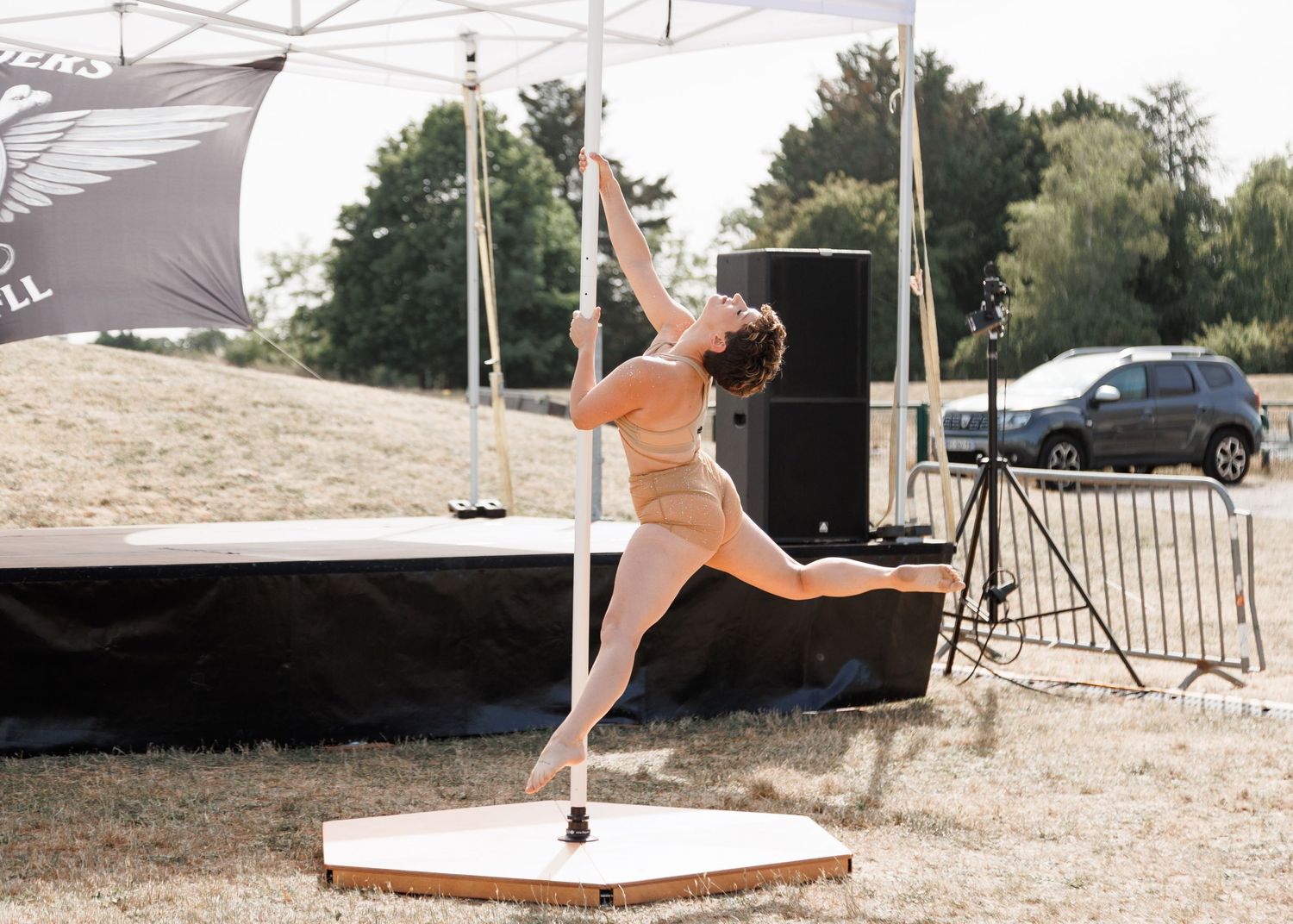 A pole dancer performs an elegant aerial pose outdoors at a festival, wearing a nude-colored outfit on a wooden stage.