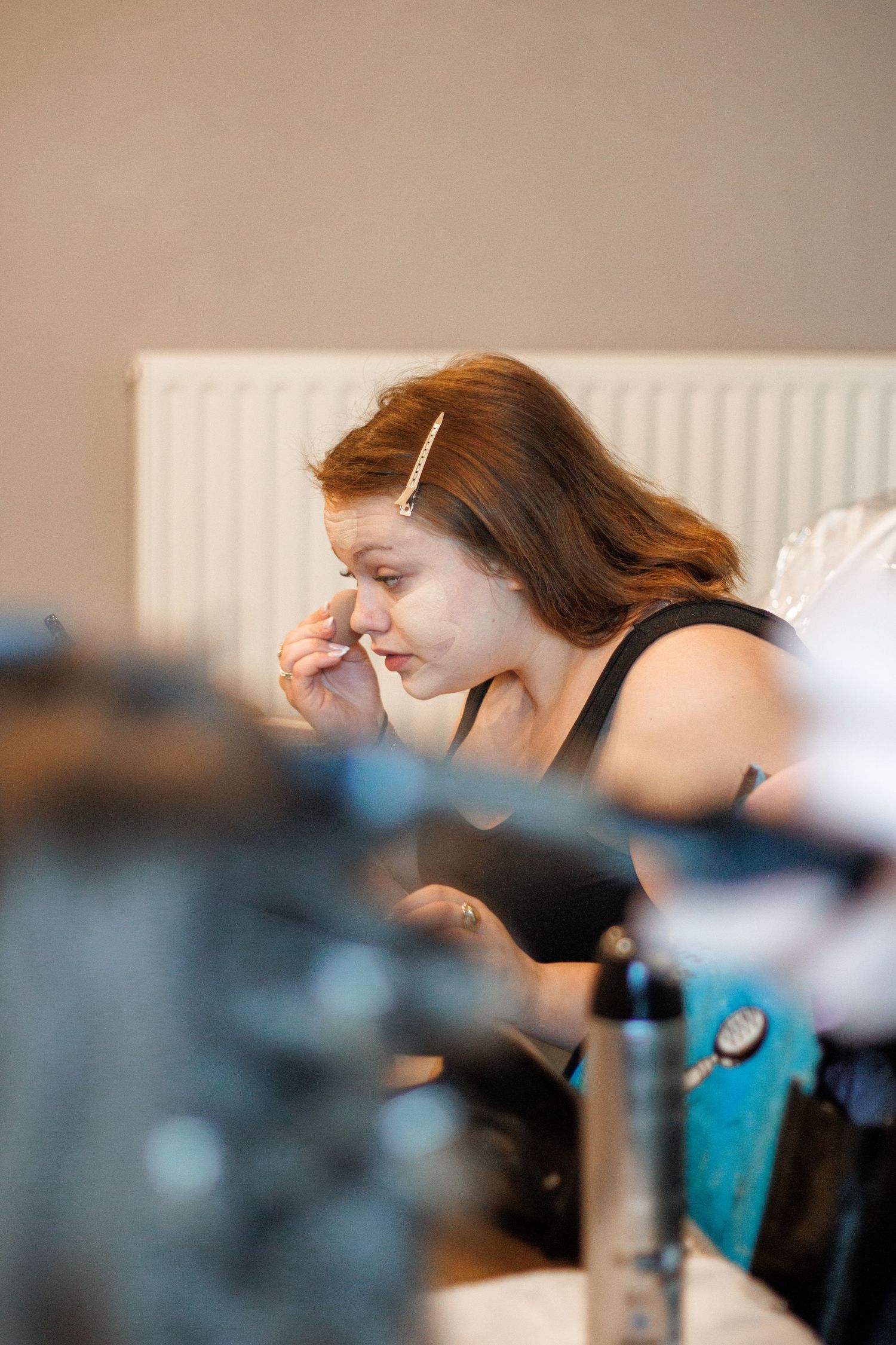 A woman applies makeup while seated at a vanity table, with a hair clip holding back her auburn hair.