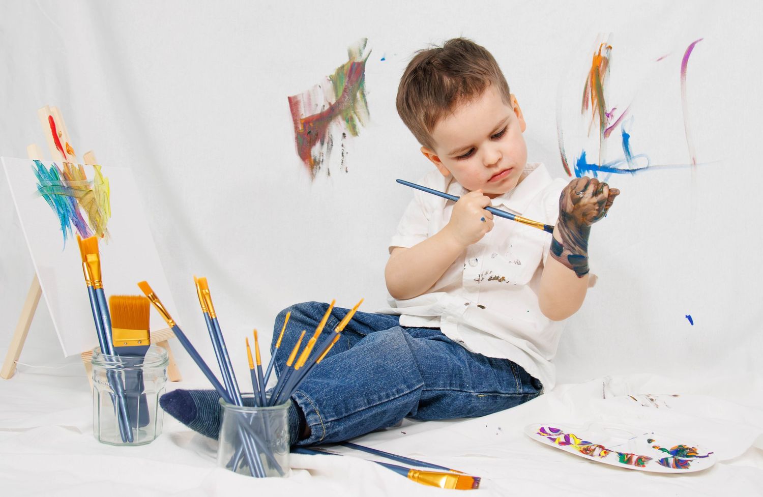 Child sitting on floor with art supplies and colorful paintings on wall.