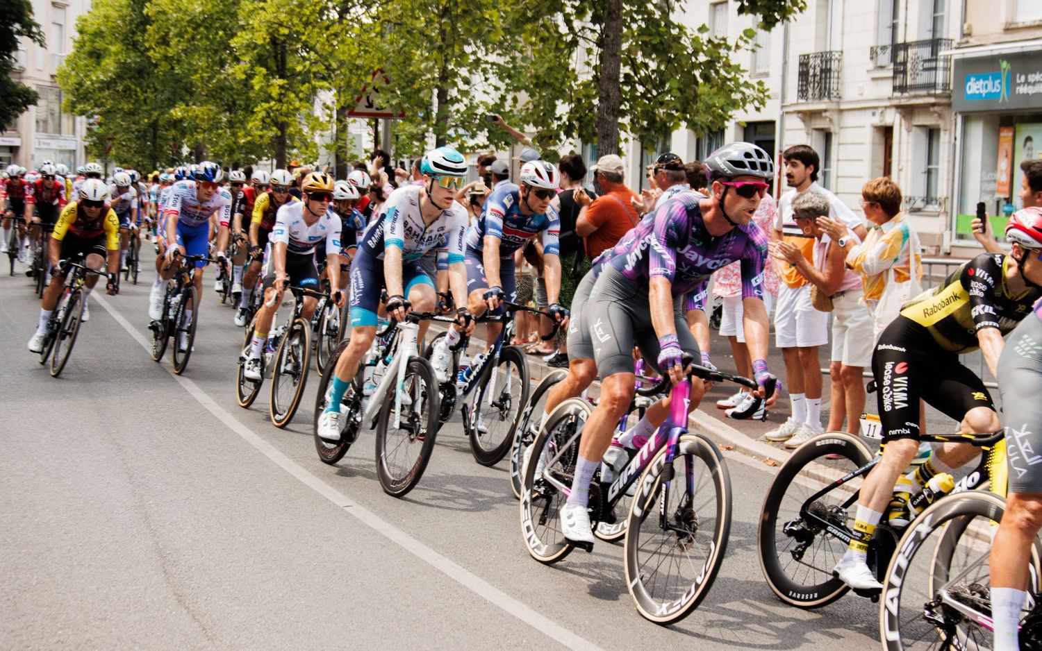Professional cyclists race in tight formation along a city street while spectators line the course.