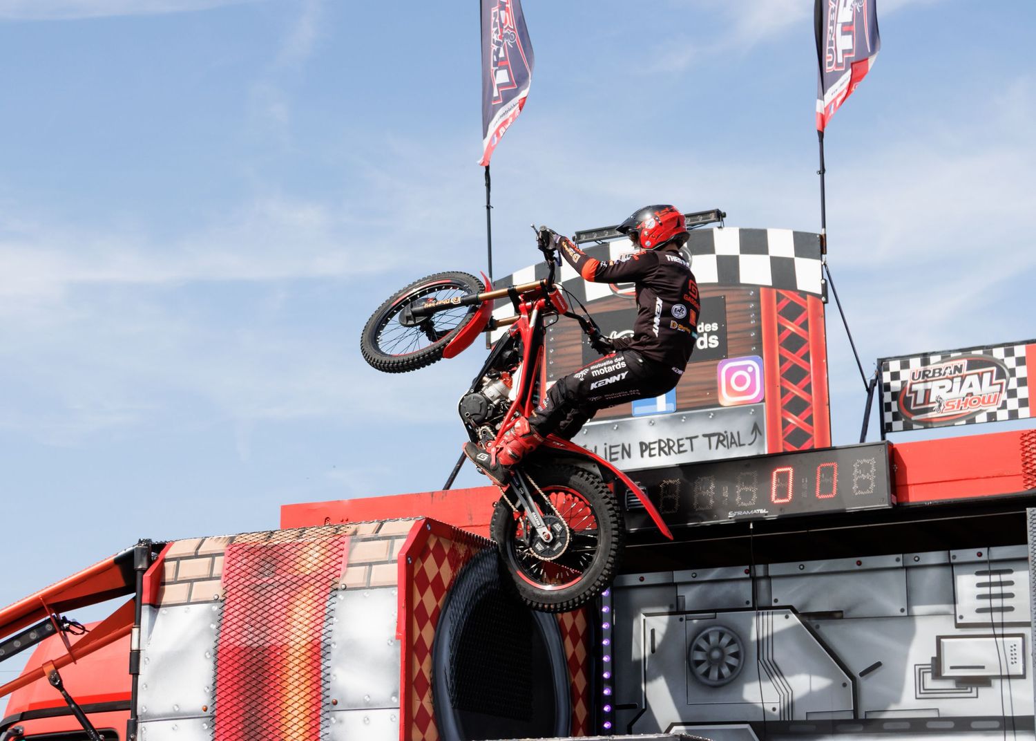 A stunt rider performs a wheelie on a motorcycle atop a red ramp at an outdoor trials event under a blue sky.