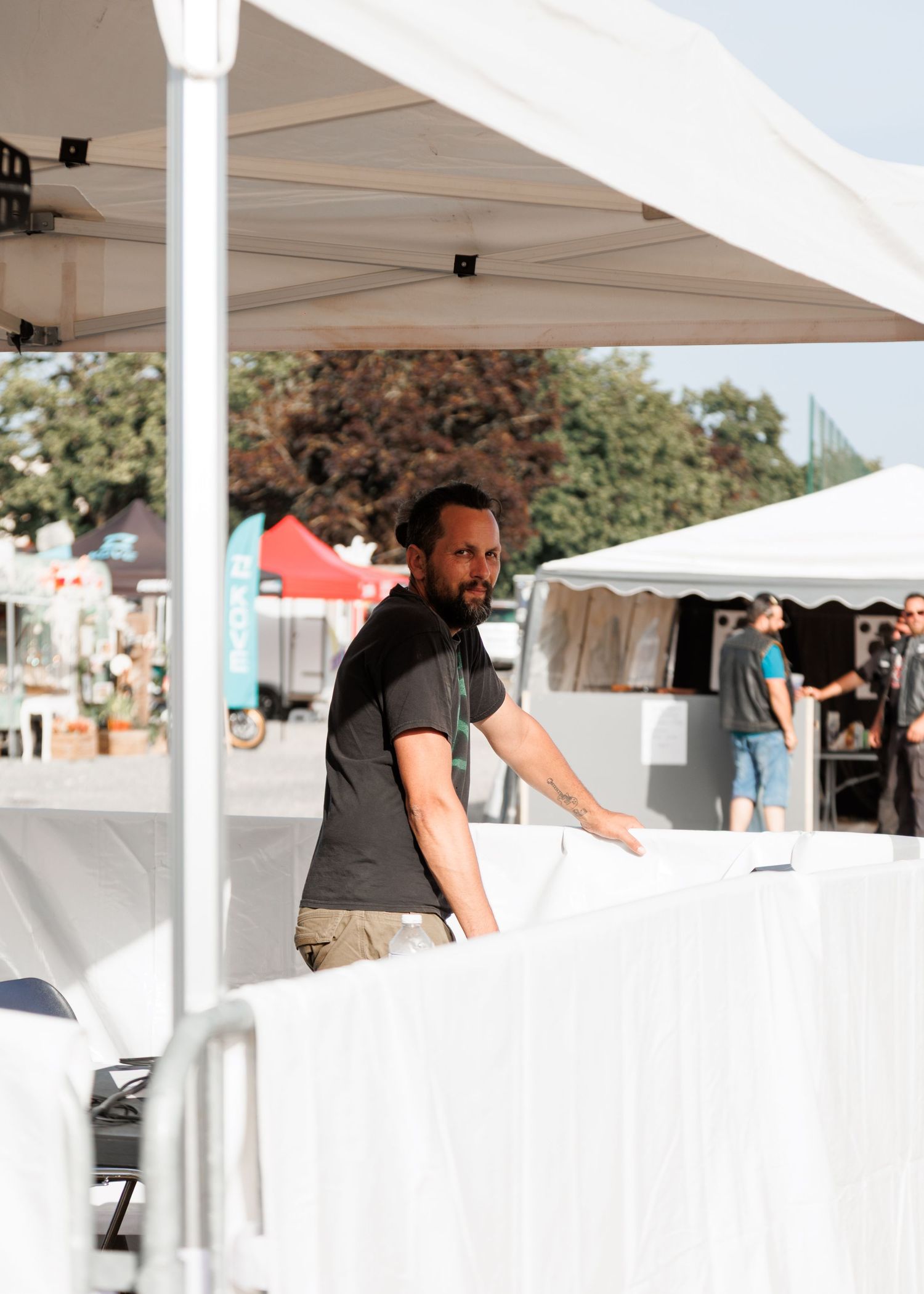 A man in a black t-shirt arranges white tables under a tent at an outdoor event on a sunny day.