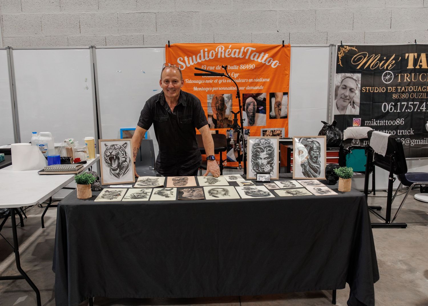Tattoo artist stands at Studio Real Tattoo booth displaying artwork prints on black draped table at convention.