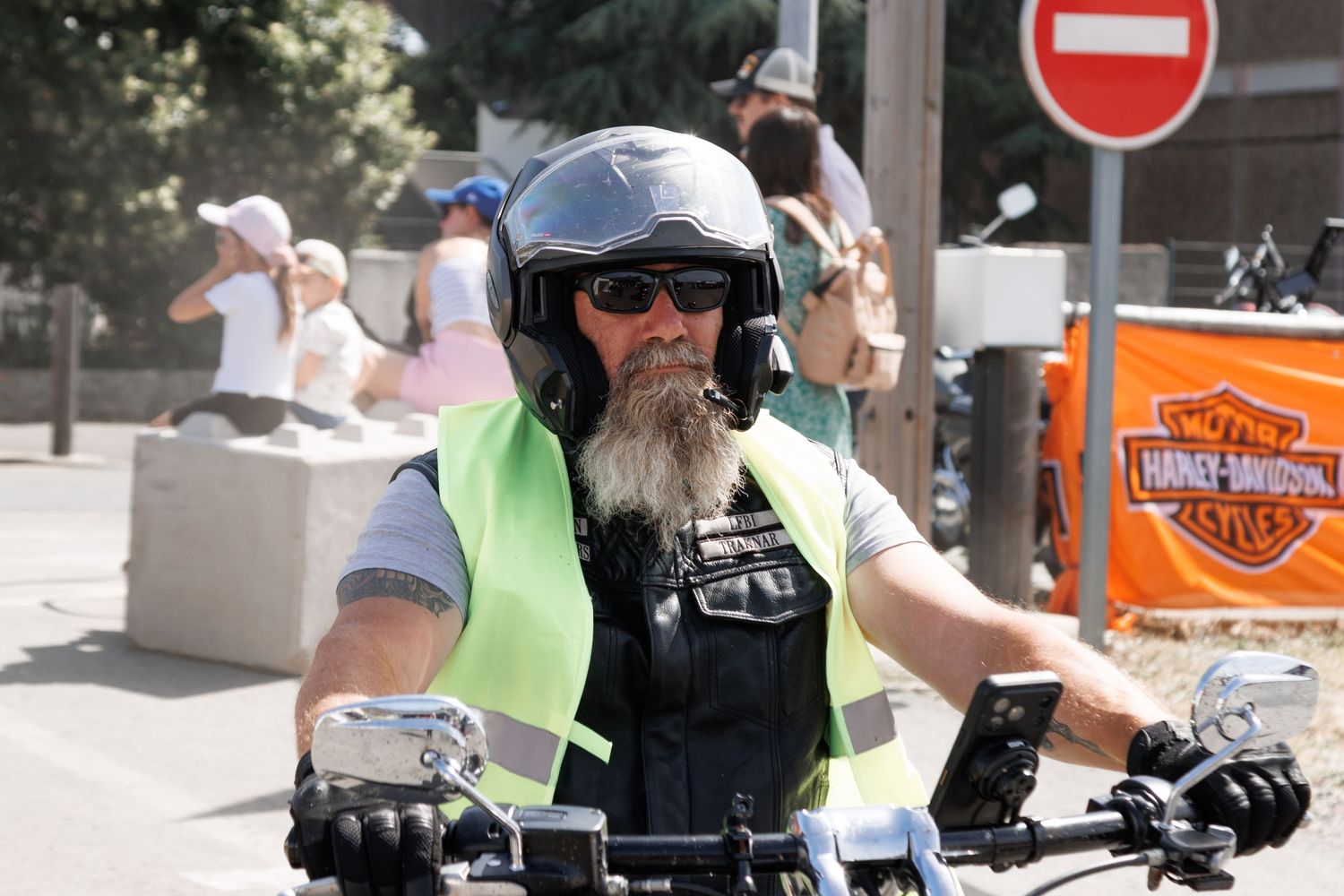 Bearded biker wearing helmet and yellow vest rides Harley-Davidson motorcycle at outdoor rally event.