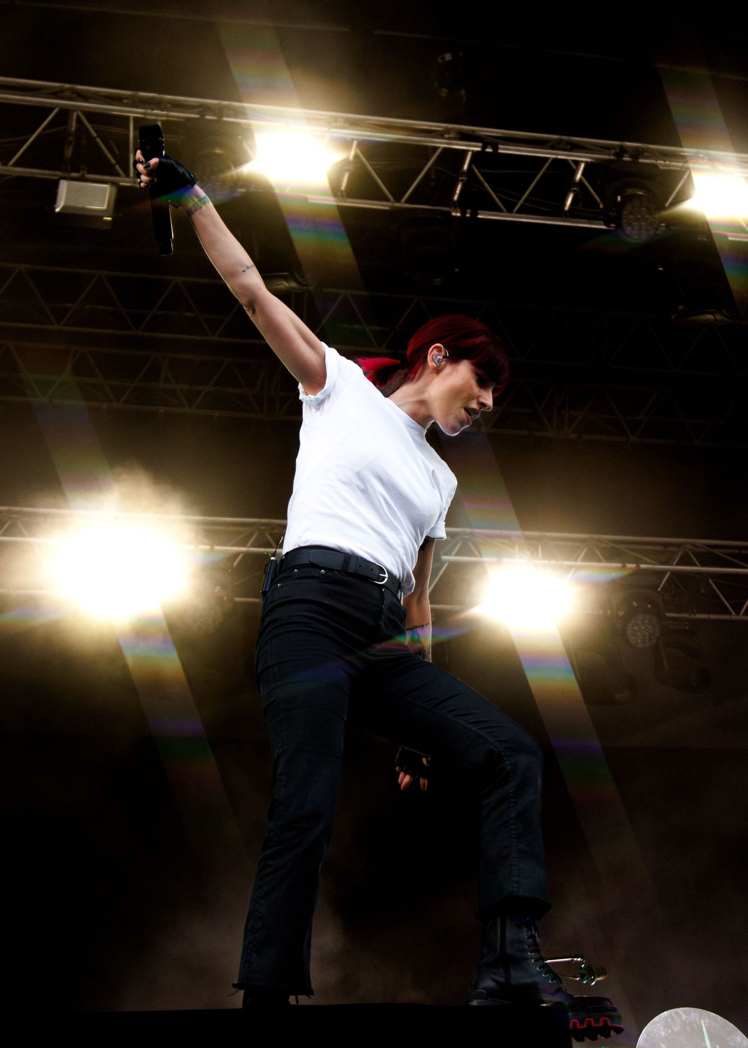 Energetic performer with red hair in white tee raises fist on stage under bright concert lights.