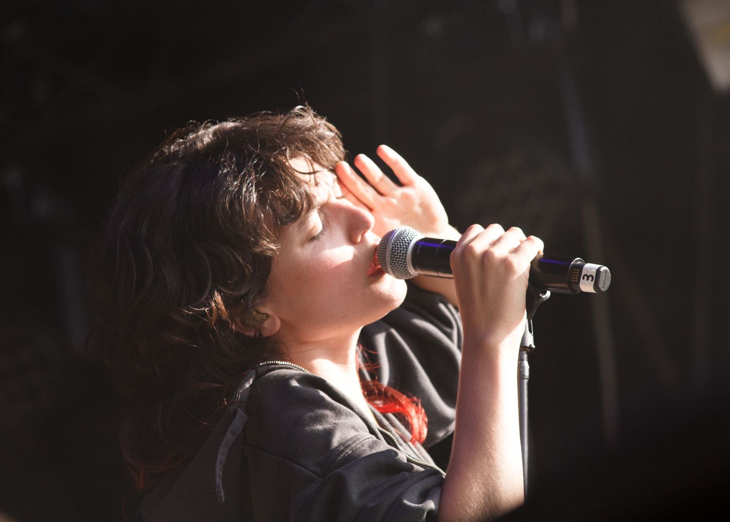 Young singer performing on stage, holding microphone close to mouth, with tousled hair and dark shirt under bright stage lighting.
