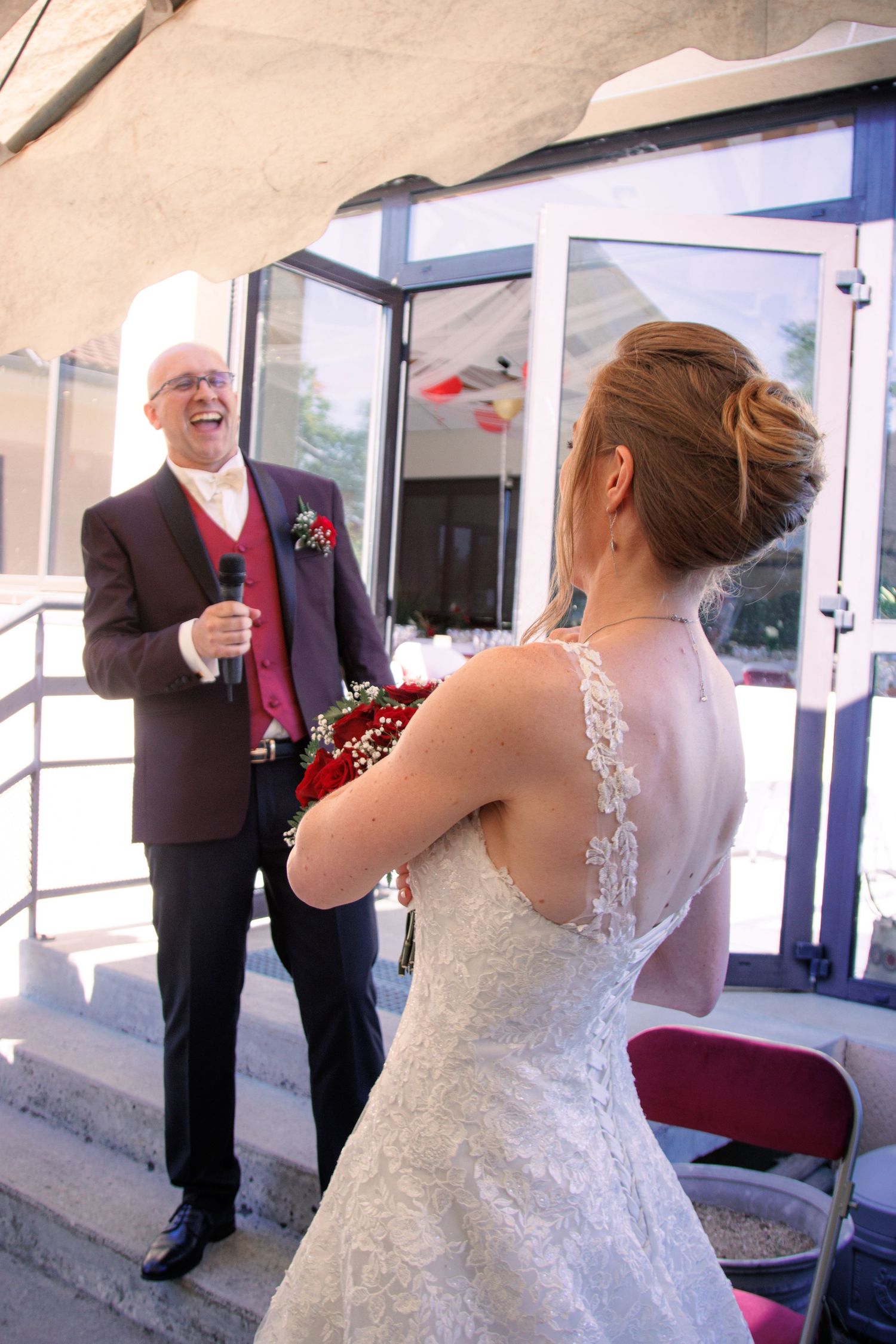 Laughing bald groom in burgundy suit speaking into microphone facing bride in lace wedding dress.