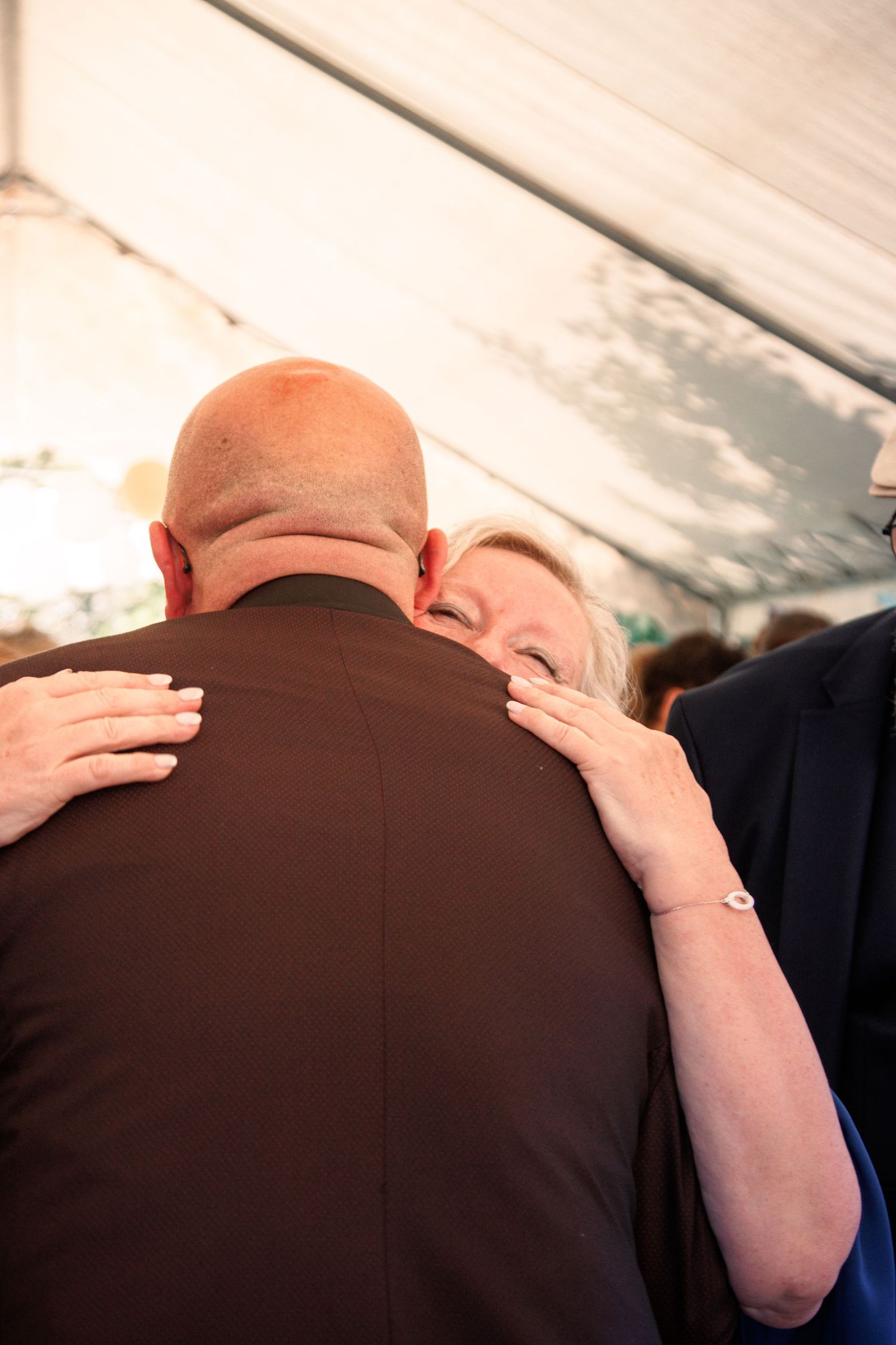 Emotional wedding moment as older woman embraces bald groom under white tent.