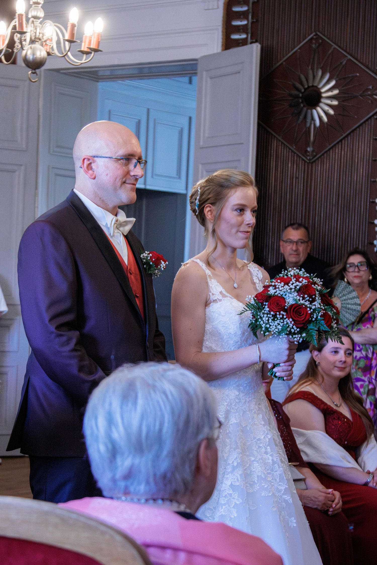 Bride holding red rose bouquet stands with bald groom in purple suit during indoor wedding ceremony with guests seated.