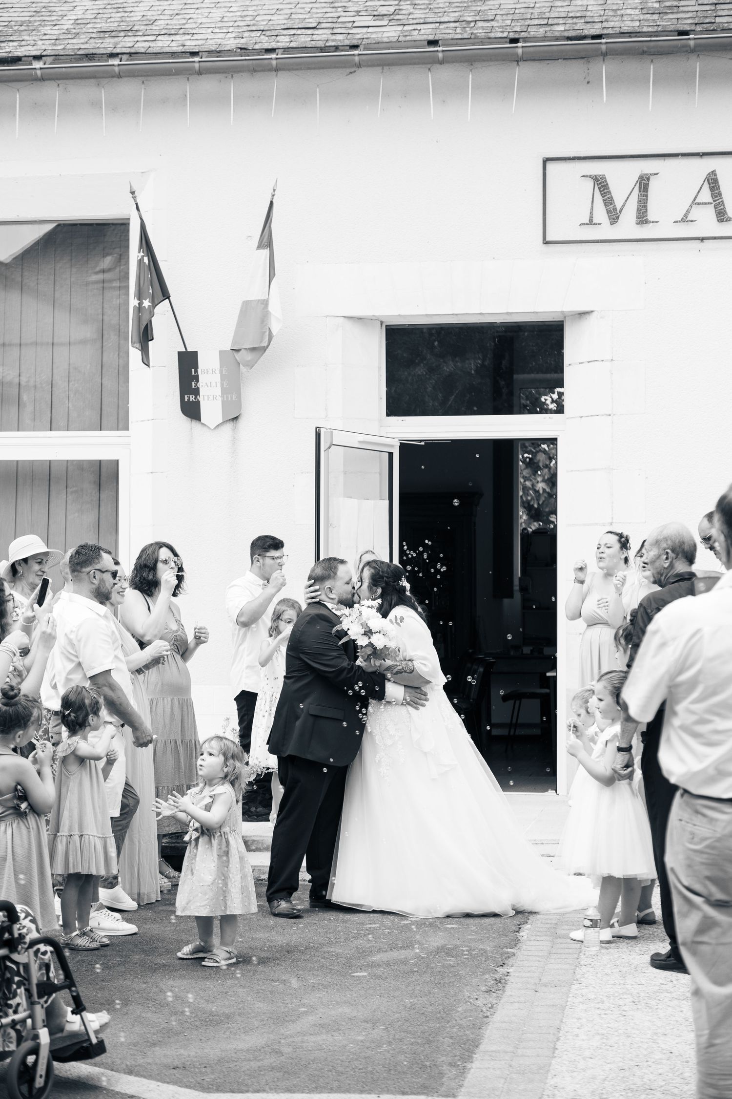 Black and white photo of a bride and groom kissing outside a French mairie surrounded by wedding guests.