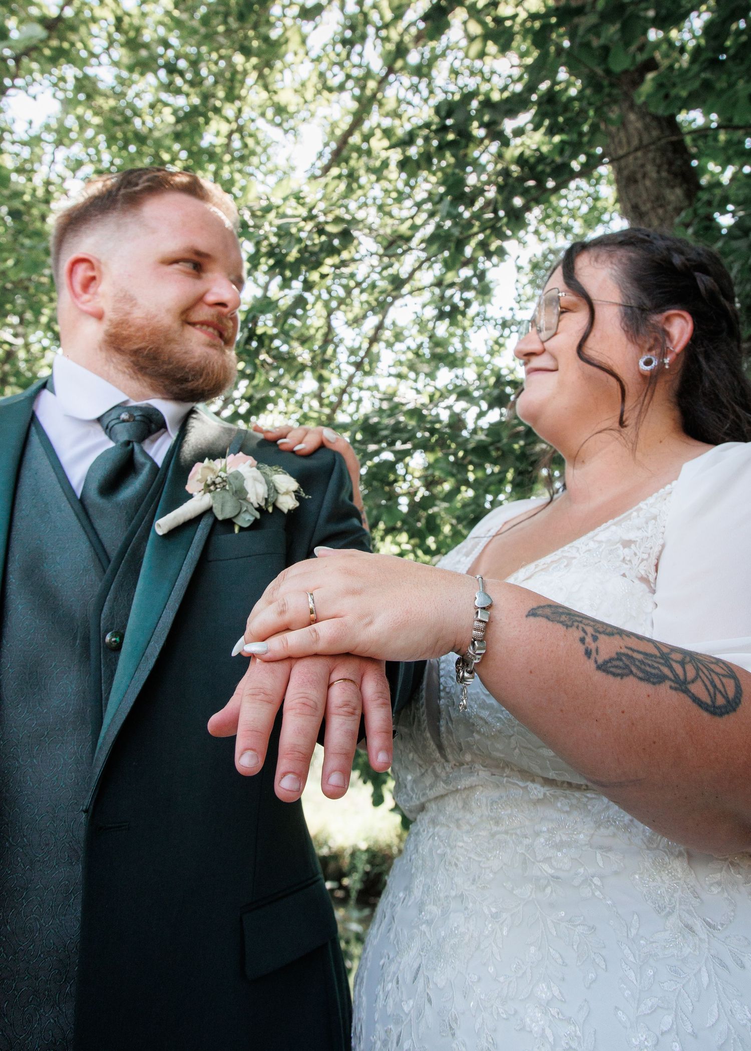 Newlyweds show off wedding rings while smiling at each other under green trees.