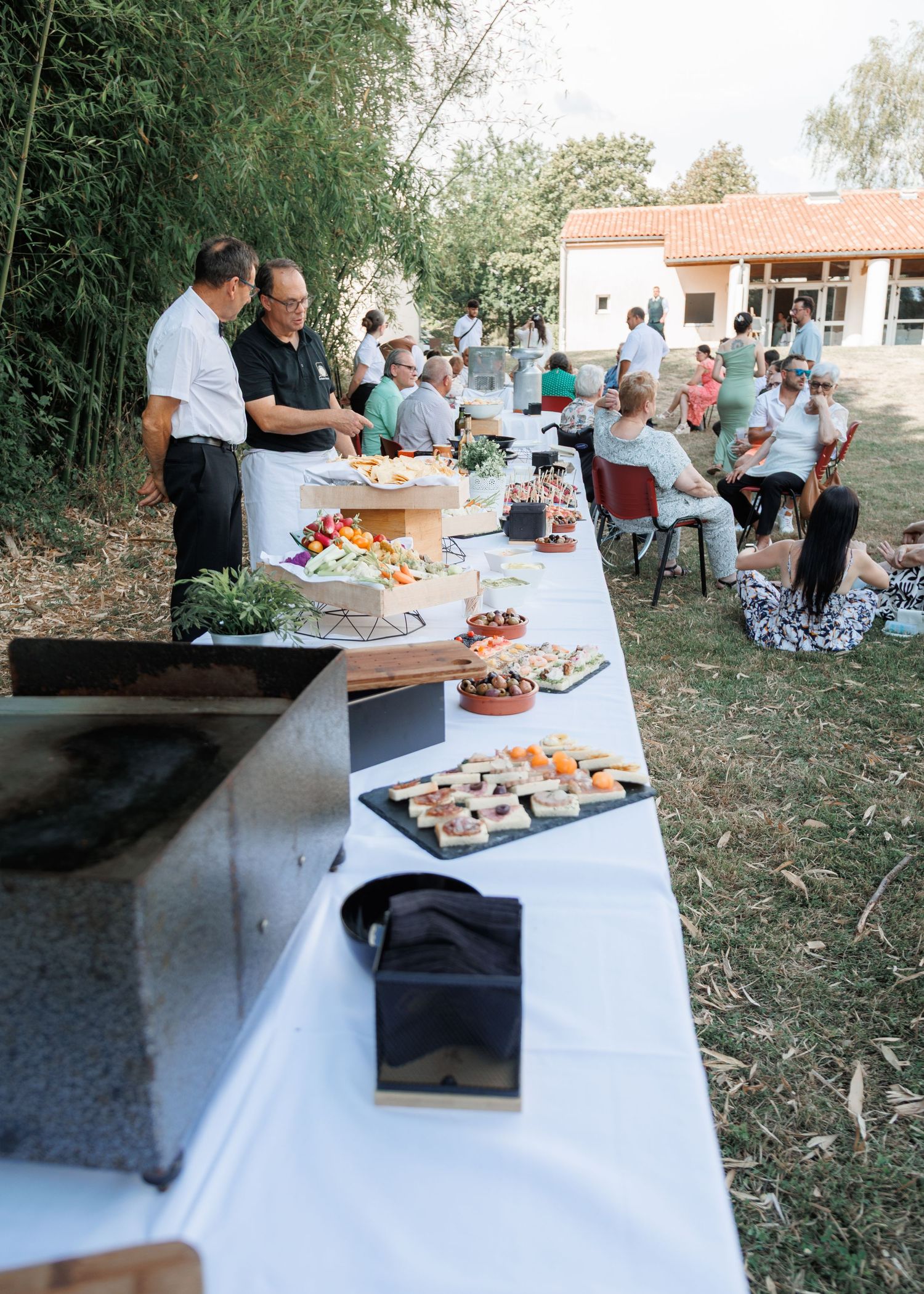 Catered outdoor wedding buffet table with assorted appetizers as guests relax in chairs nearby.