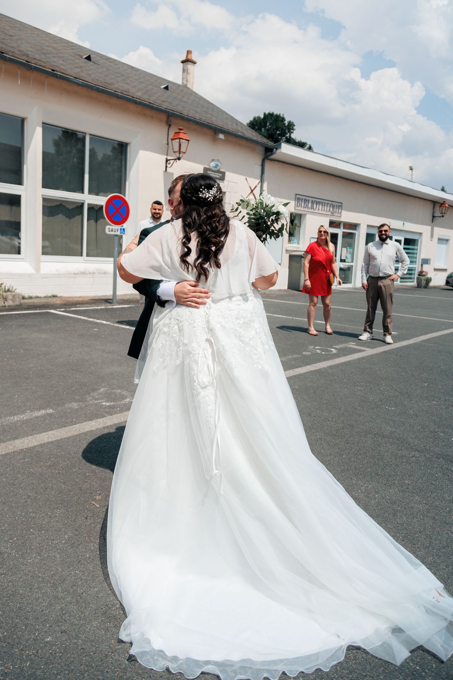 Bride in elegant white gown with long train embraced from behind in outdoor wedding venue parking lot.