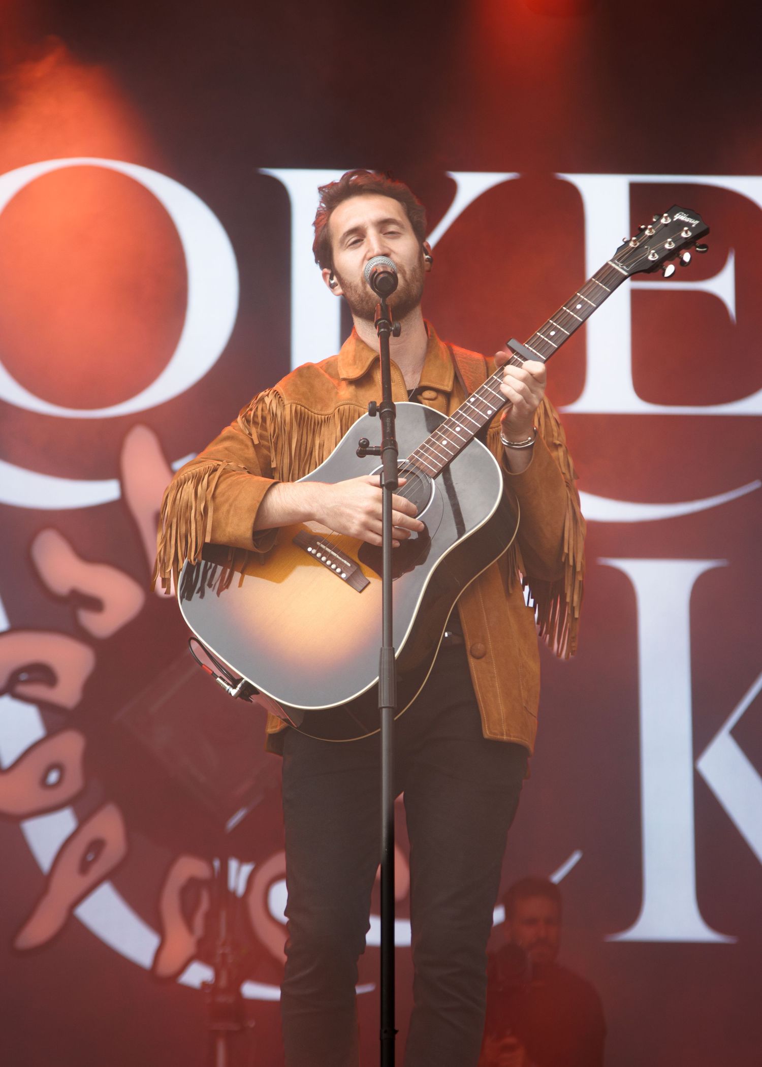 A musician in a brown fringe jacket performs on stage with a guitar under dramatic red lighting.