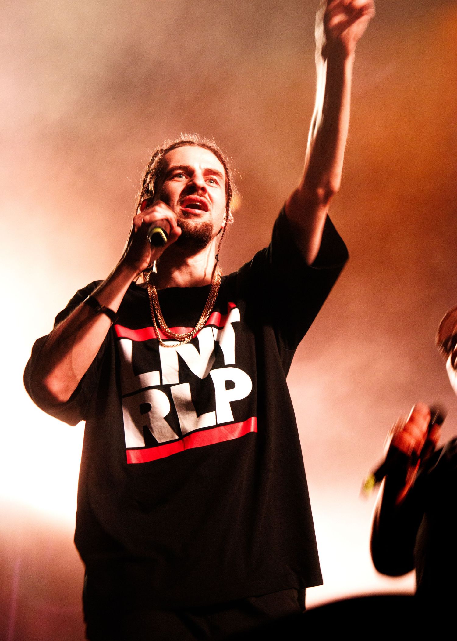 Performer in black shirt with white text sings passionately on stage with dramatic orange lighting in background.