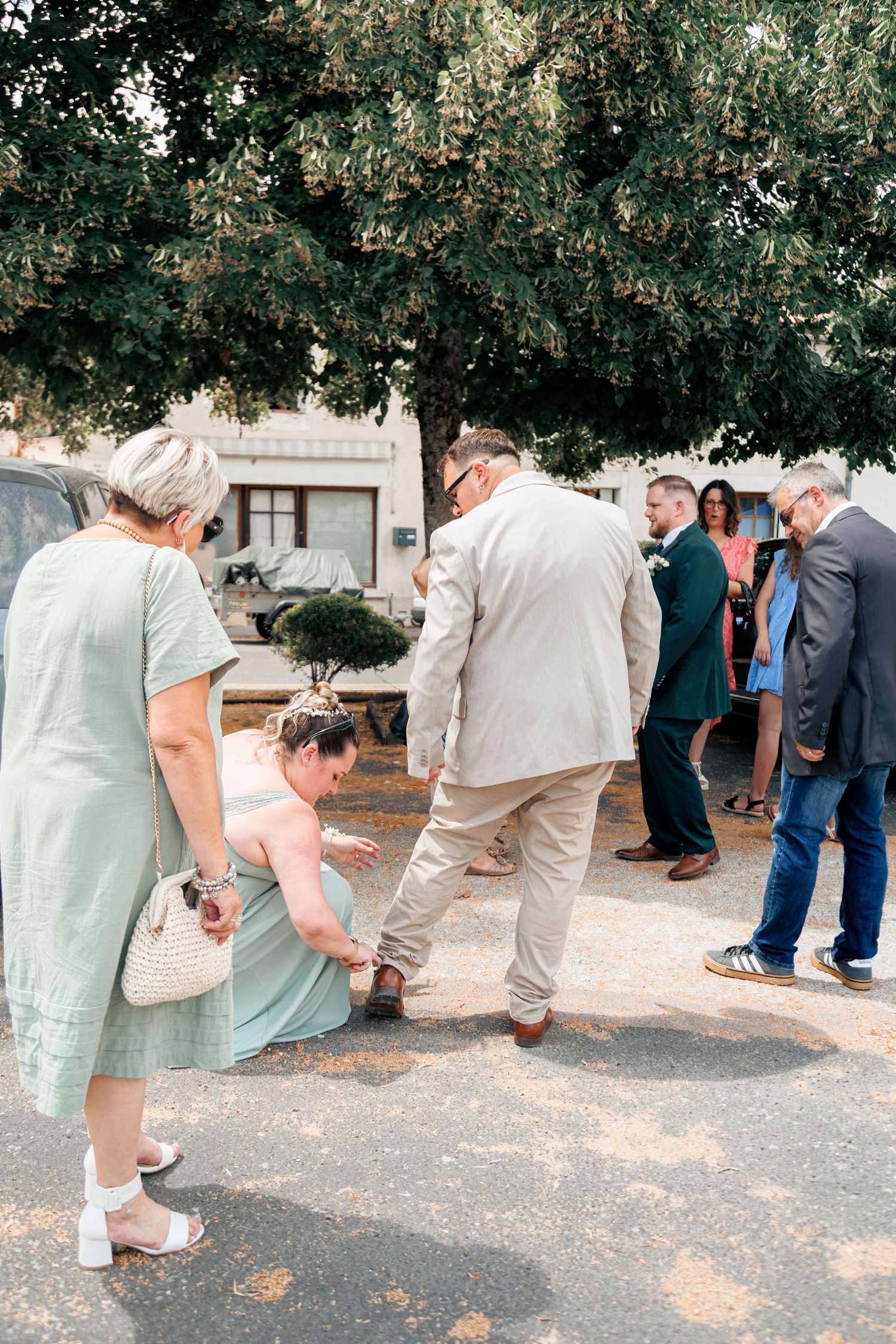 Wedding guests gather outdoors under a large tree, with one woman crouching to help another guest near the ground.