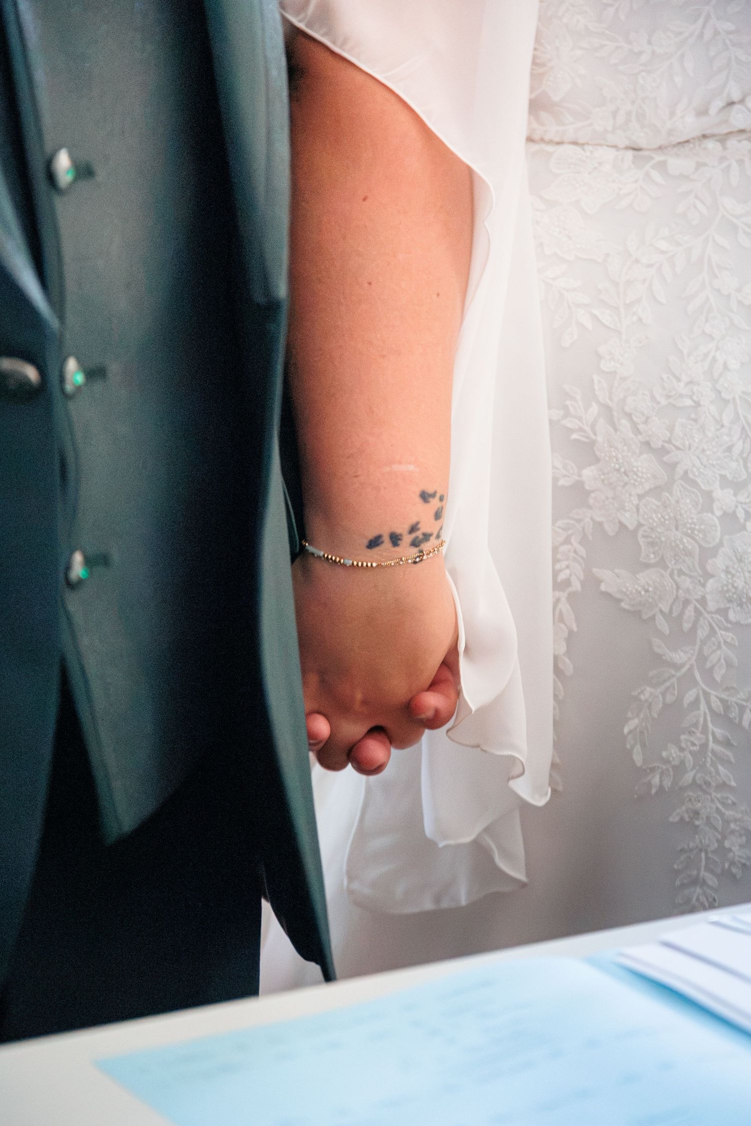 Bride and groom holding hands at wedding ceremony, bride wearing white dress with floral tattoo and delicate bracelet.